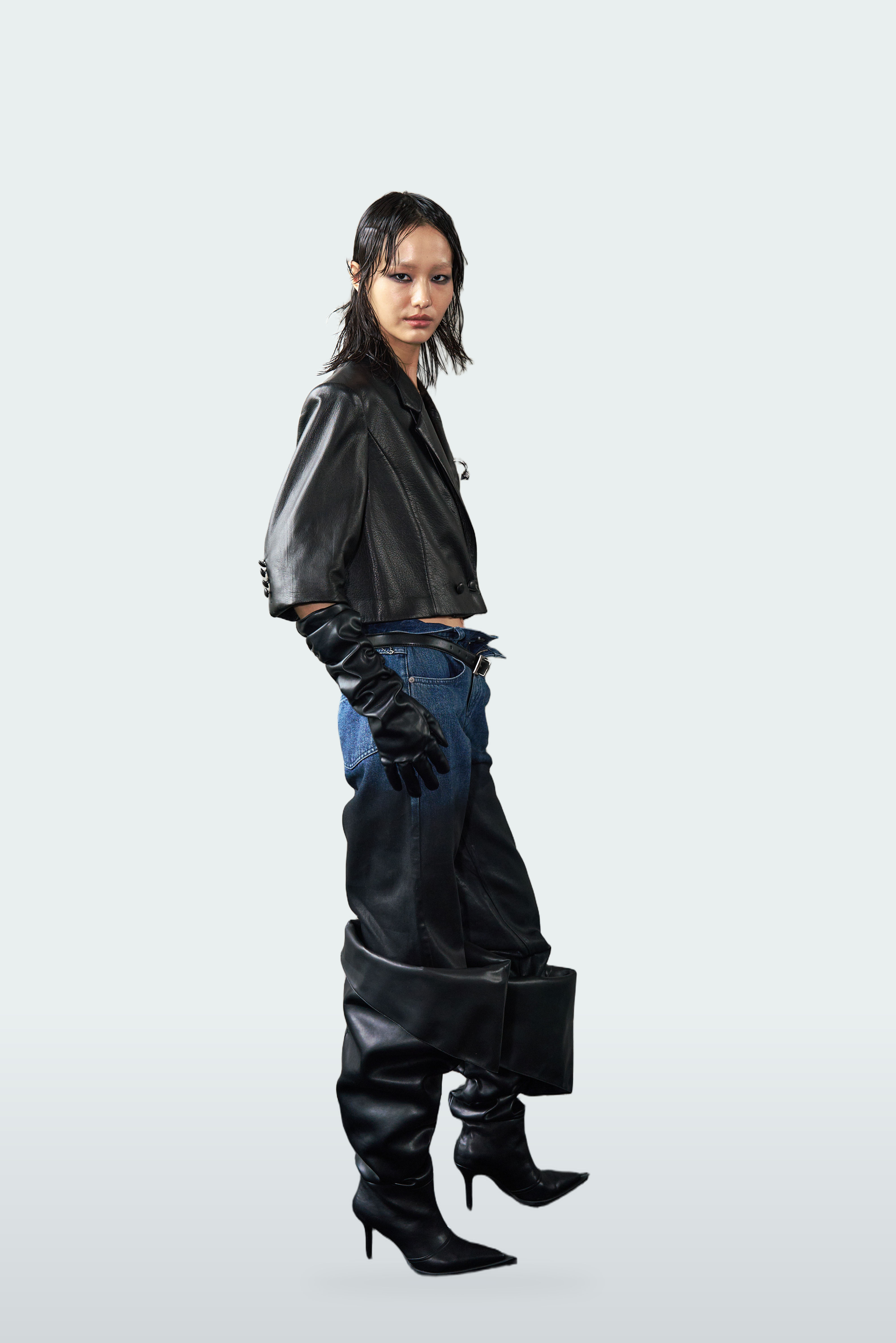 A model is shown in profile wearing a black leather cropped jacket, blue jeans, black leather gloves and boots, holding a black rectangular box against a light gray studio background.