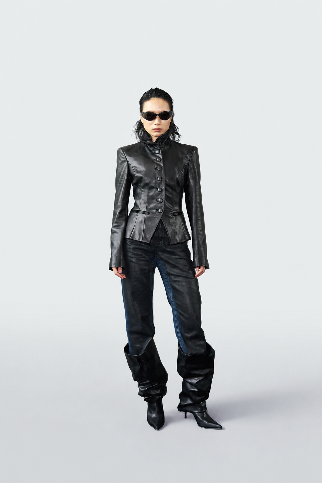 A model stands against a light gray studio background wearing a black leather jacket with structured shoulders, black jeans, and black pointed-toe heeled boots.