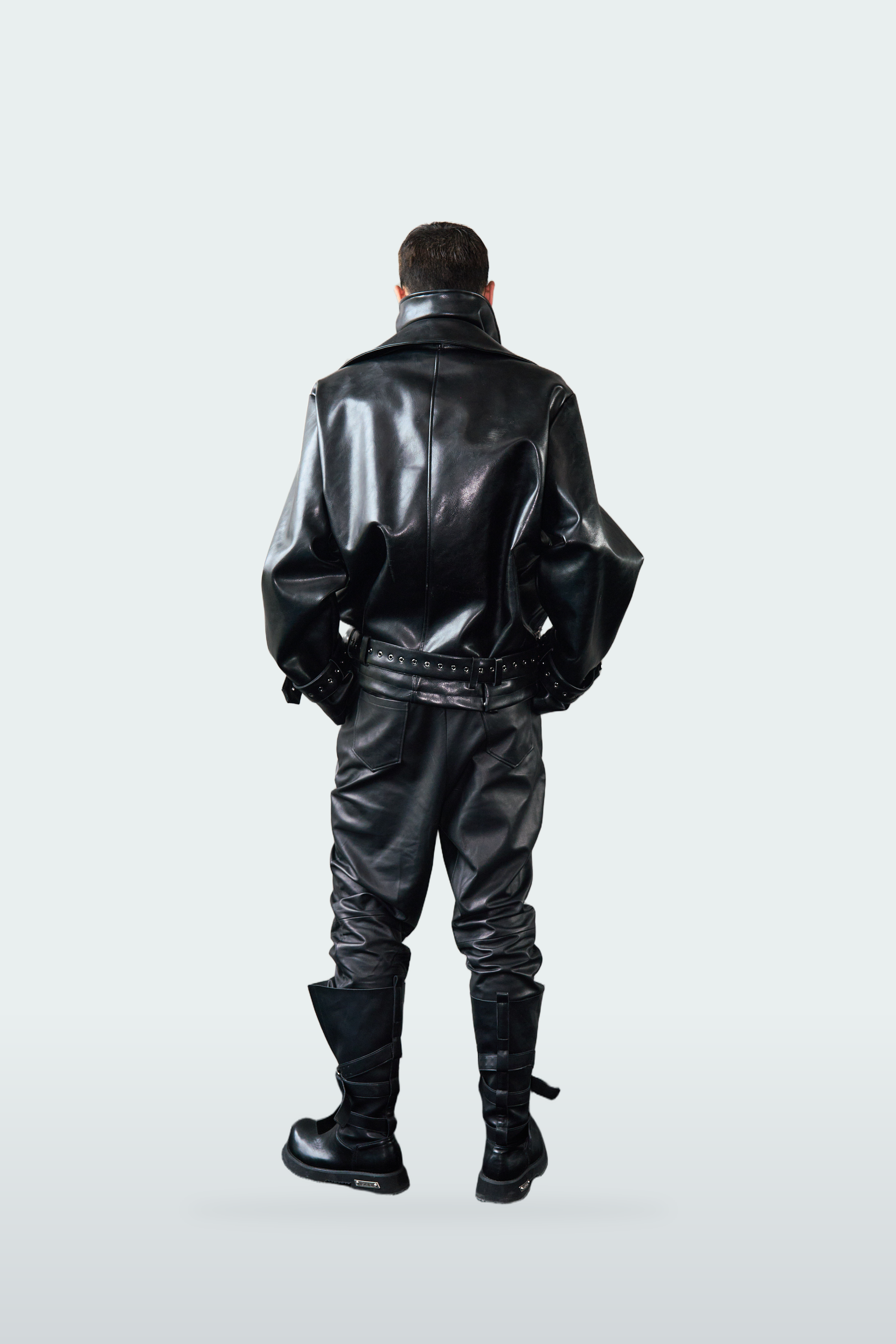 A model viewed from behind wearing a shiny black faux-leather jacket, matching black pants, and black boots against a light gray studio background.