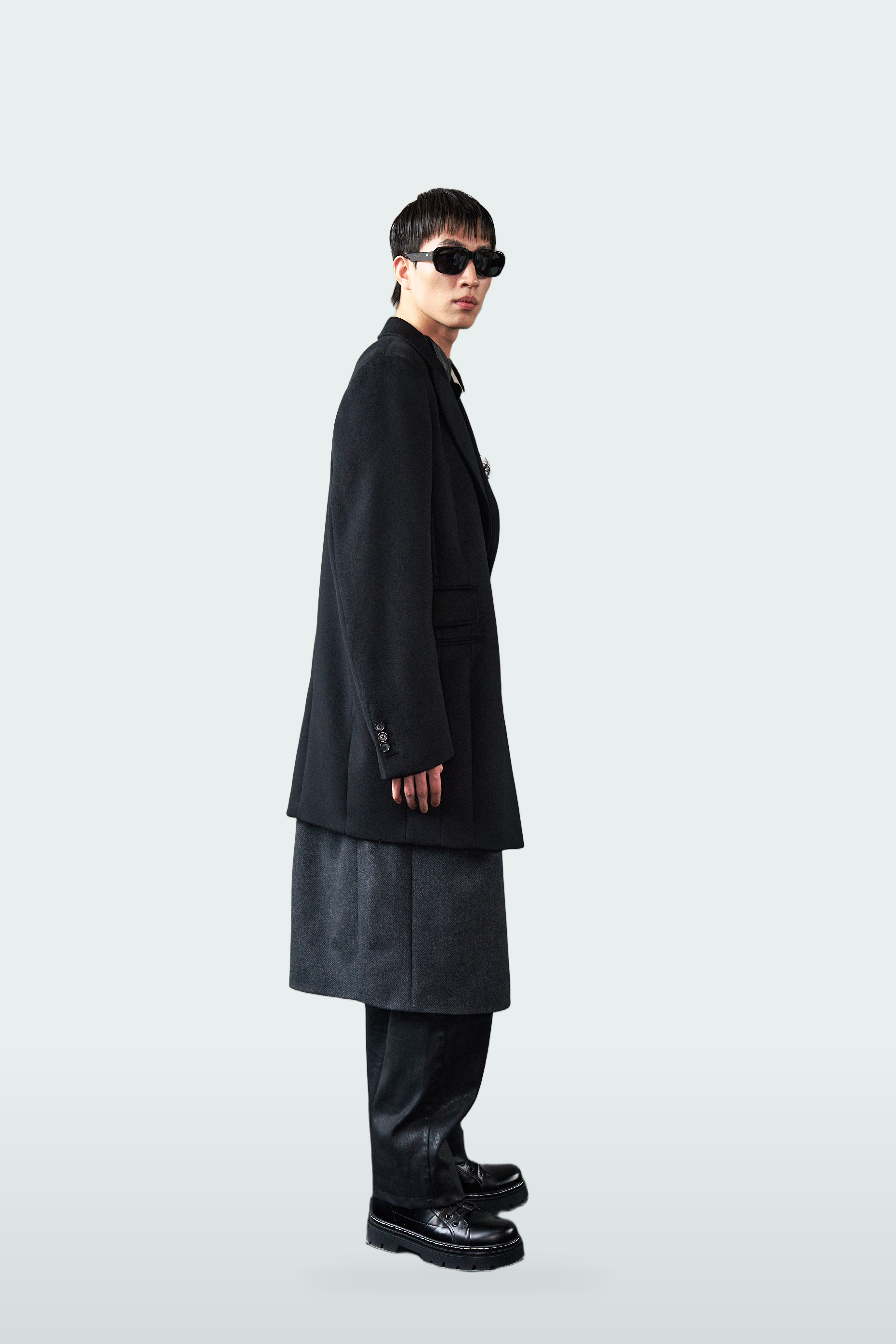 A model in side profile wearing a long black blazer, a dark gray pleated skirt, black trousers, and black platform sunglasses against a light gray studio background.