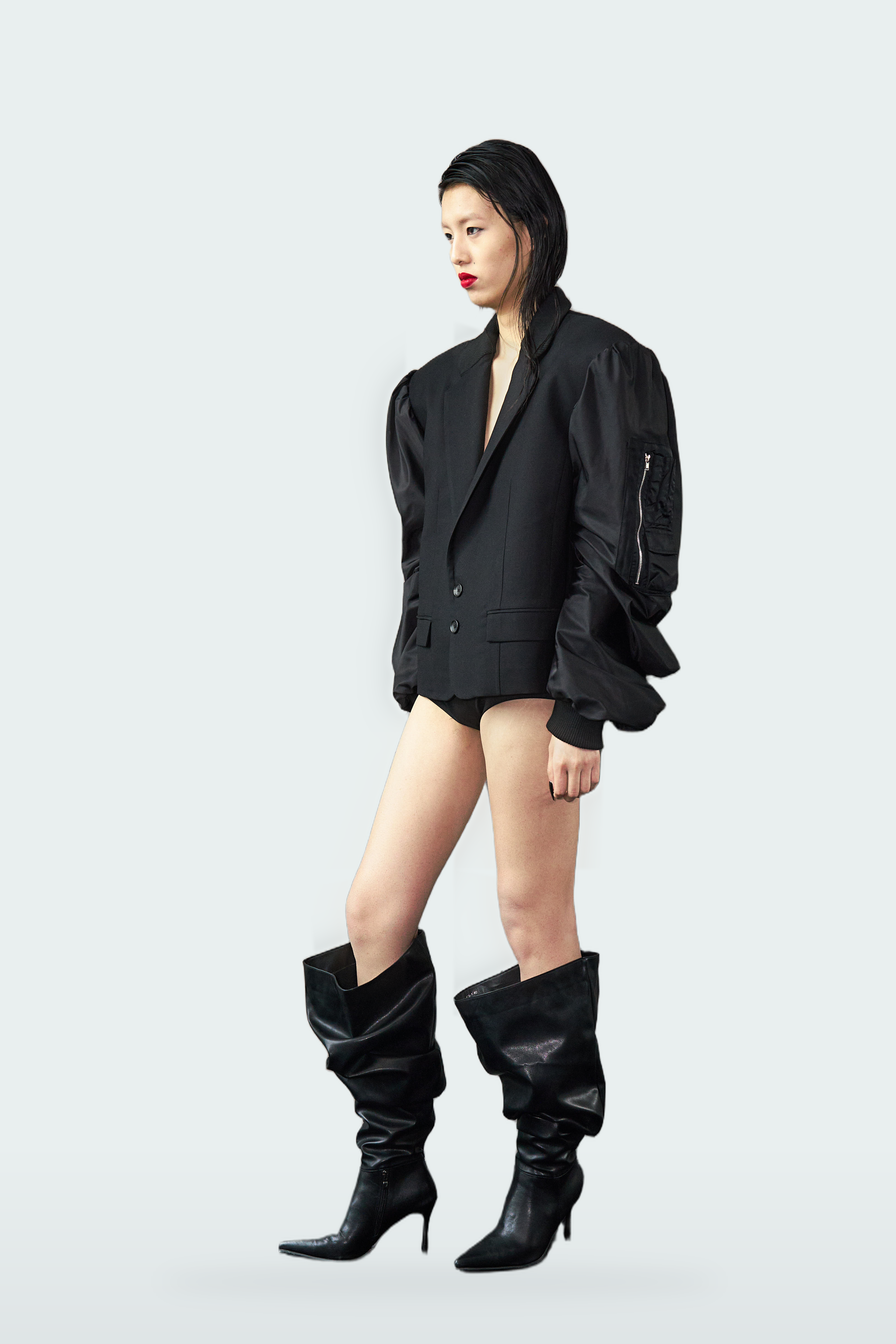 A model wearing a black short-sleeve blazer with dramatic ruffled sleeves, matching black shorts, and black knee-high heeled boots, posed against a light gray studio background.