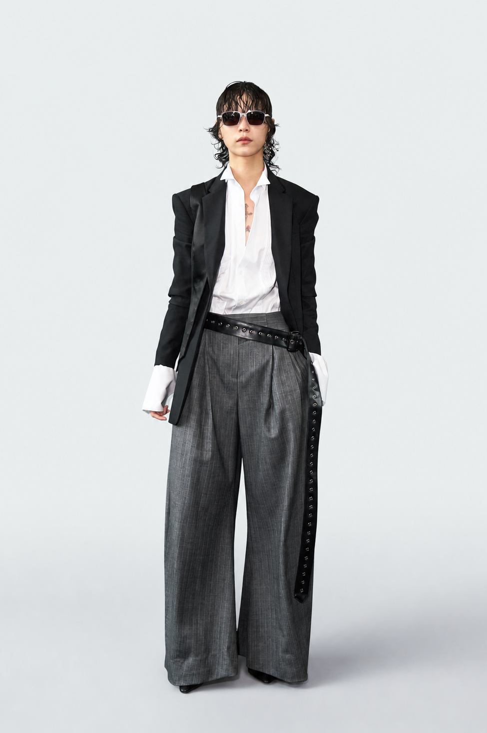 A model stands against a light gray studio background wearing a black blazer over a white shirt, paired with wide-leg gray trousers featuring a black side panel with silver studs, a black belt, and dark sunglasses.