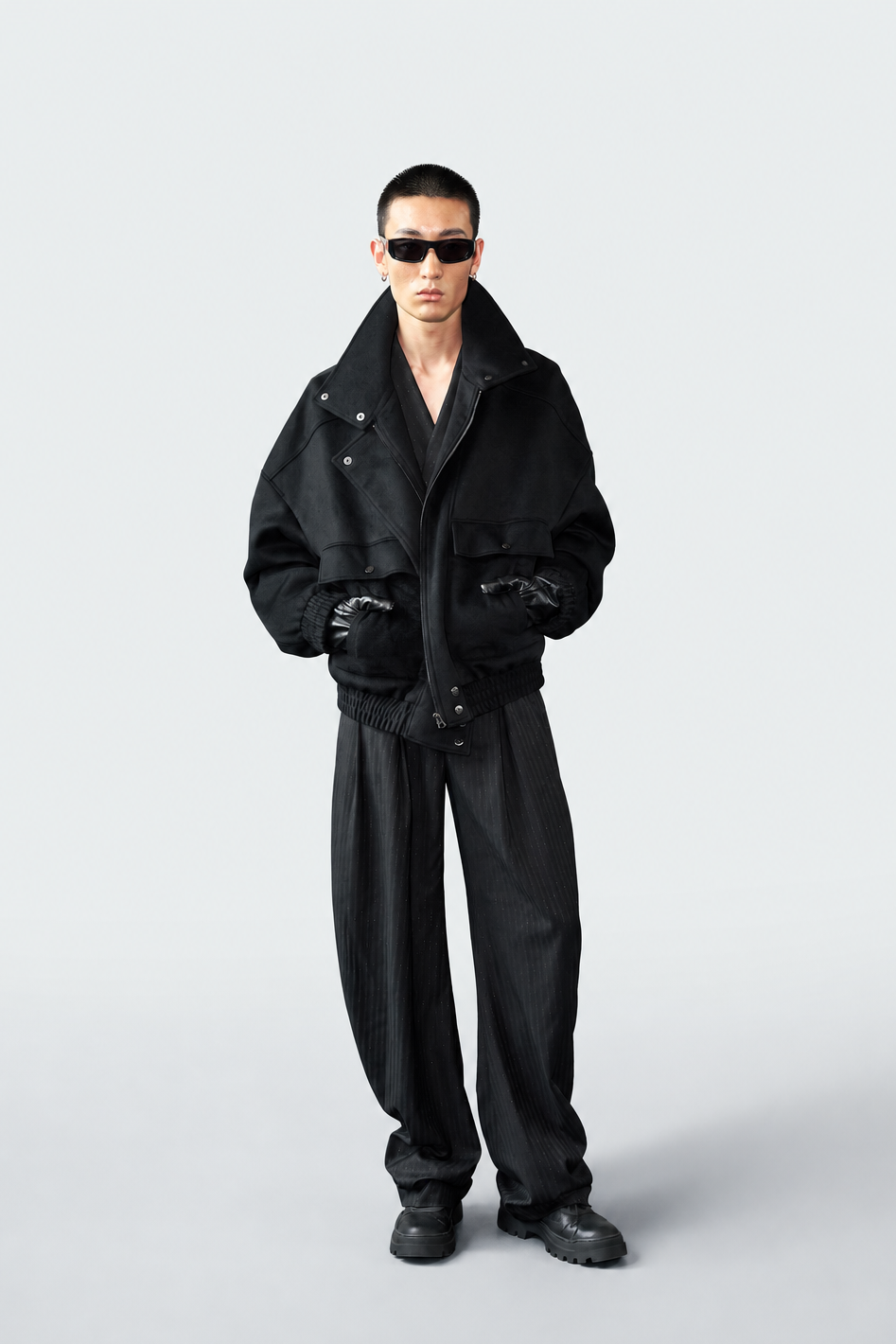 A model wearing a black oversized jacket, matching black trousers, black gloves, and black sunglasses, standing against a light gray studio background.