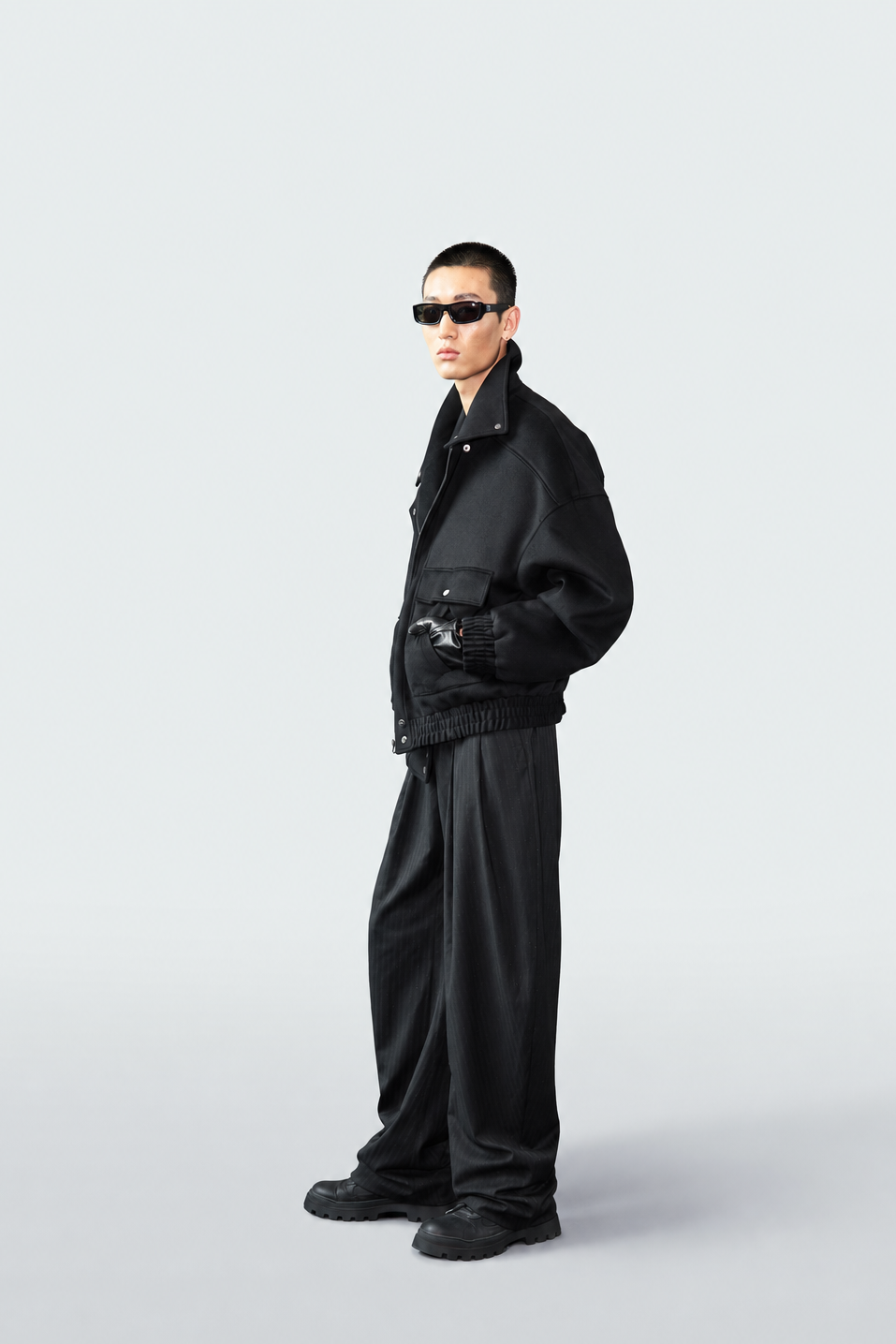 A model wearing a black oversized jacket, matching black trousers, black sunglasses, and black shoes, standing in profile against a light gray studio background.