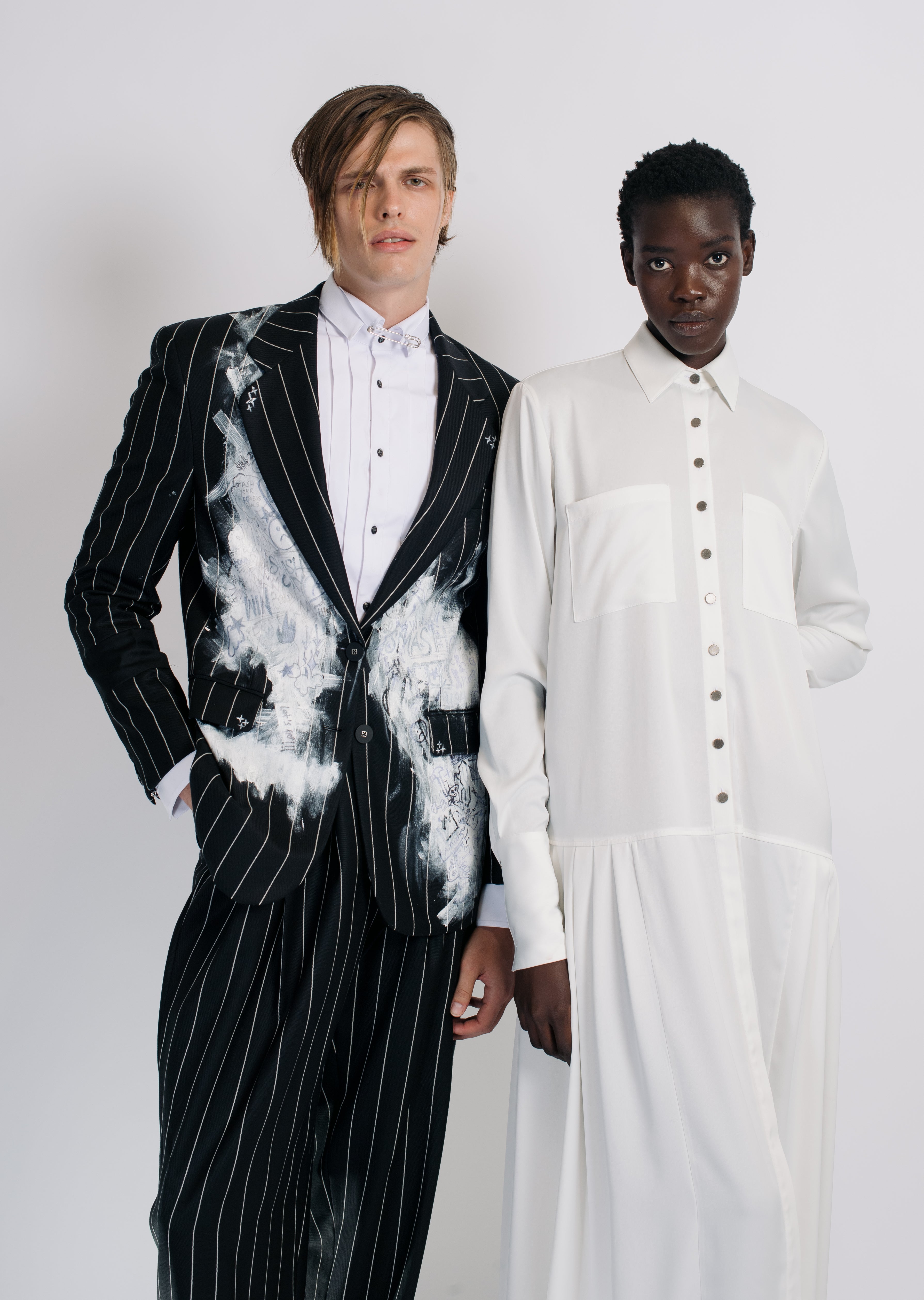Male model wearing White Pleated Tuxedo Shirt under a black pinstripe blazer with artistic white brushstroke accents, standing beside a model in a minimalist white dress, showcasing SHAO New York bespoke tailoring and refined luxury fashion.