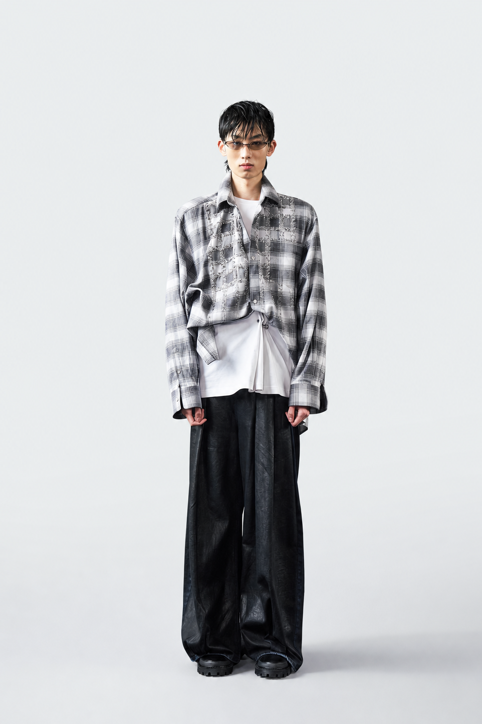 A model stands against a light gray studio background wearing a cropped black and white plaid overshirt over a white shirt, paired with loose black trousers.