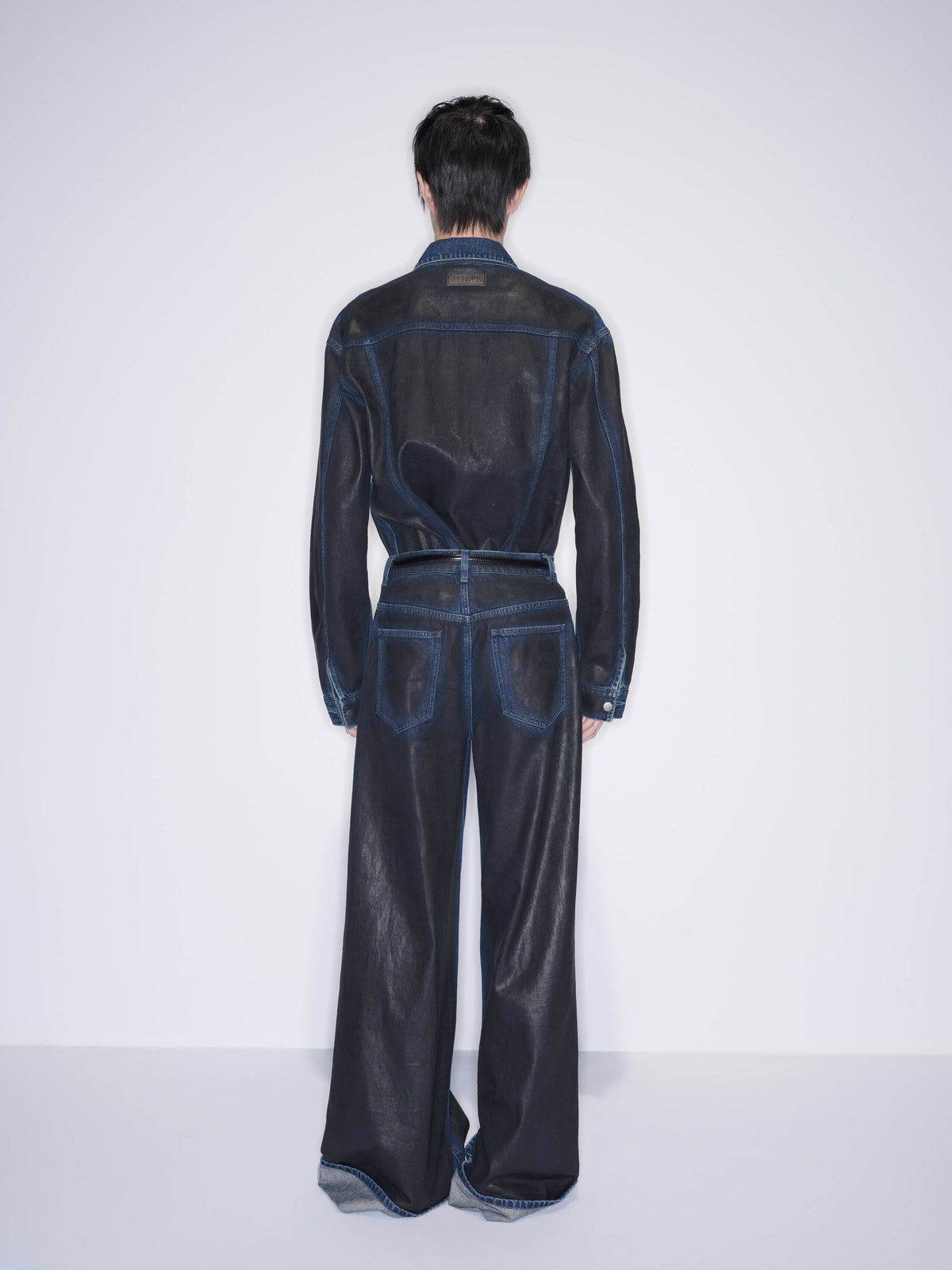 A model viewed from the back wearing a dark denim jumpsuit with a sheer mesh back panel and wide-leg trousers, set against a plain white studio background.
