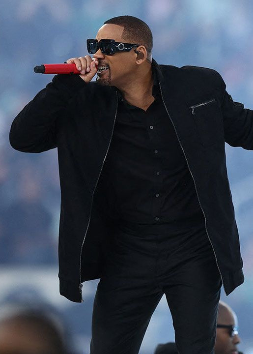 A man dressed in an all-black outfit with a zippered jacket and sunglasses performs energetically while holding a red microphone against a blurred outdoor background.
