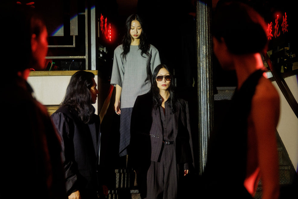 A woman in a black suit and sunglasses stands on a staircase in a dimly lit, atmospheric setting, surrounded by other people in dark clothing.