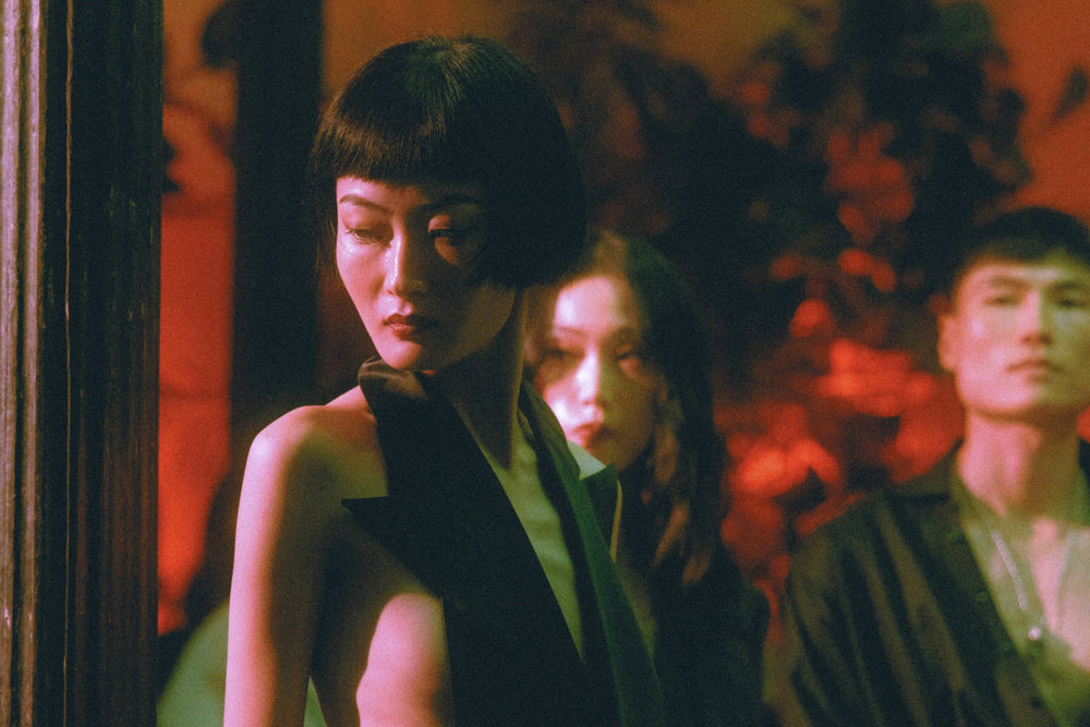 A moody, stylized photograph featuring a woman in the foreground wearing a black sleeveless top, with two other women blurred in the background under warm, red-orange ambient lighting.