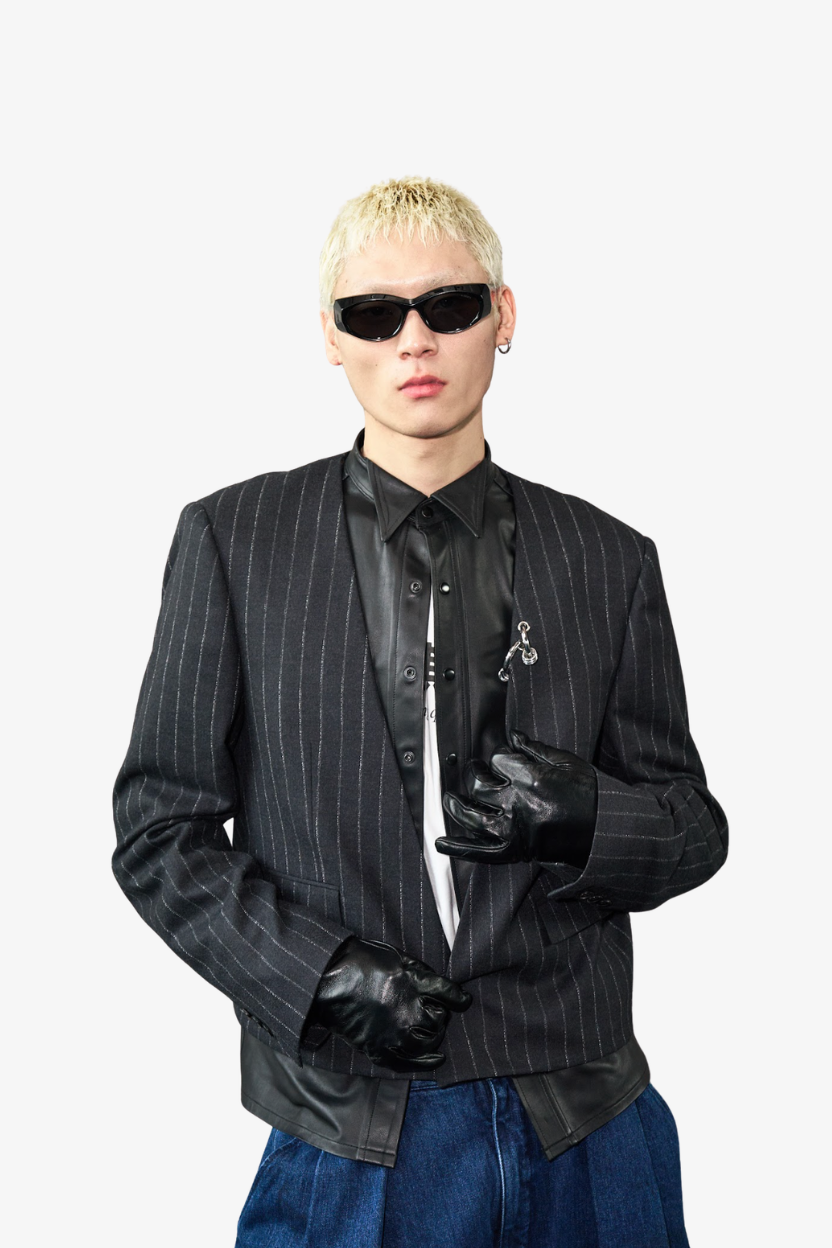 A model with short blonde hair wearing a black pinstripe blazer, black leather shirt, black gloves, and black sunglasses, paired with blue jeans against a light gray background.