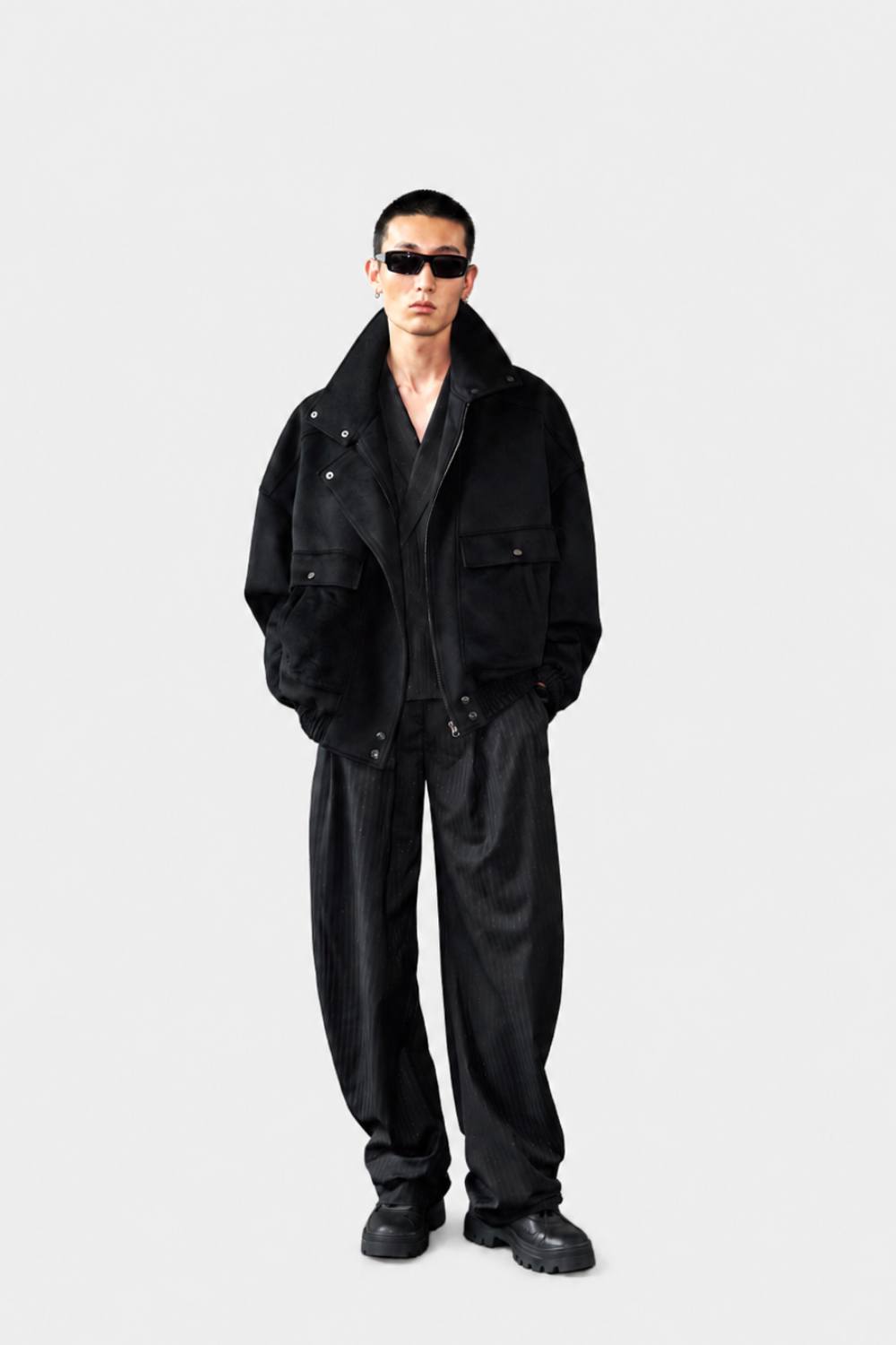 A model wearing a black corduroy jacket, black corduroy trousers, black sunglasses, and black boots, standing against a light gray studio background.