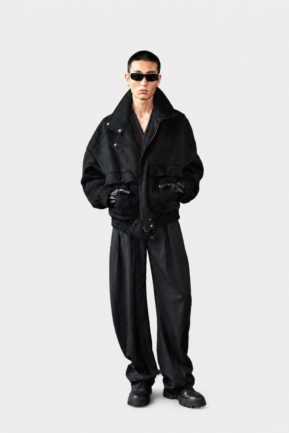 A model wearing a black oversized jacket with a high collar, black pants, and black sunglasses, standing against a light gray studio background.