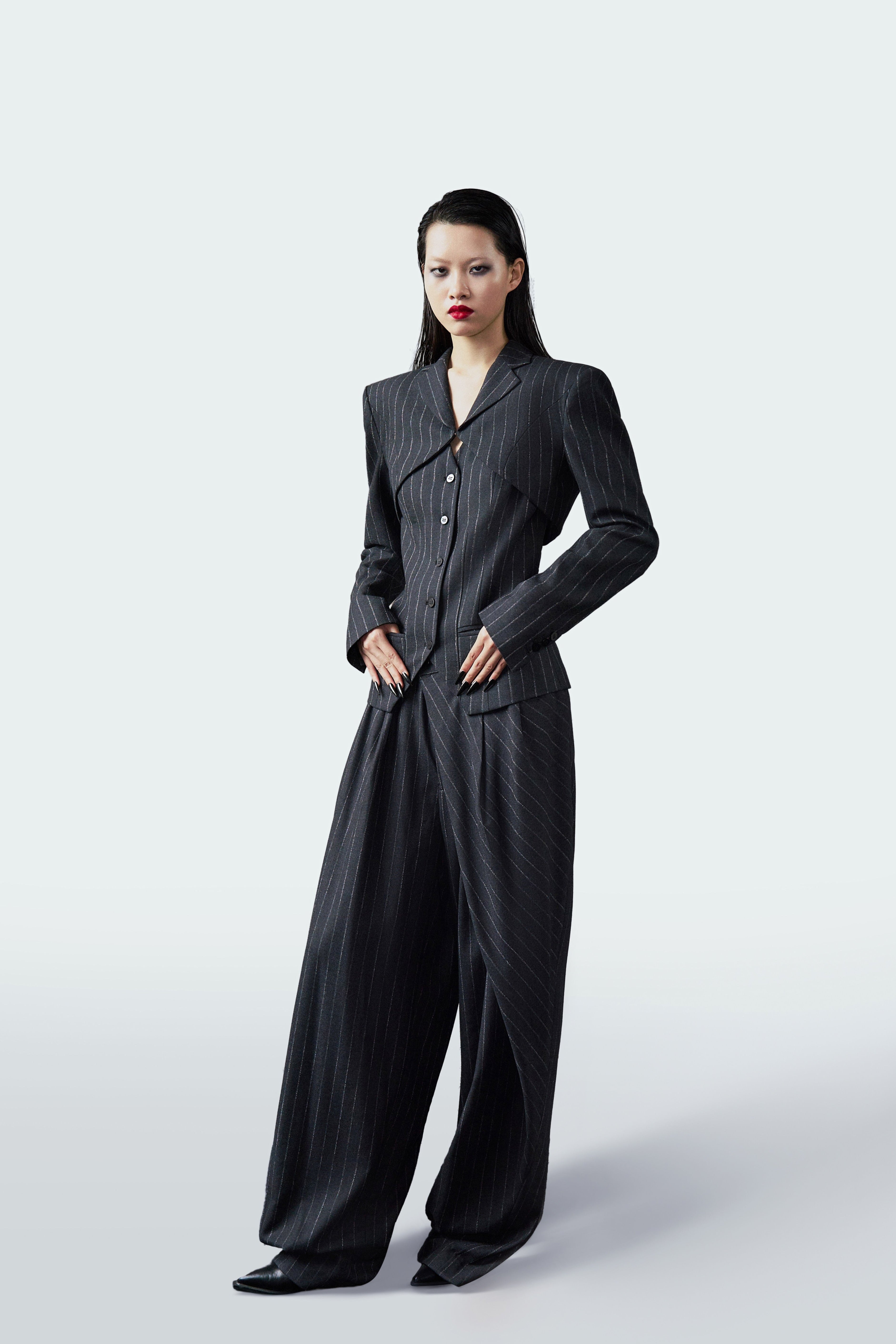 A model wearing a dark gray pinstriped jumpsuit with structured shoulders and wide-leg trousers, standing against a light gray studio background.