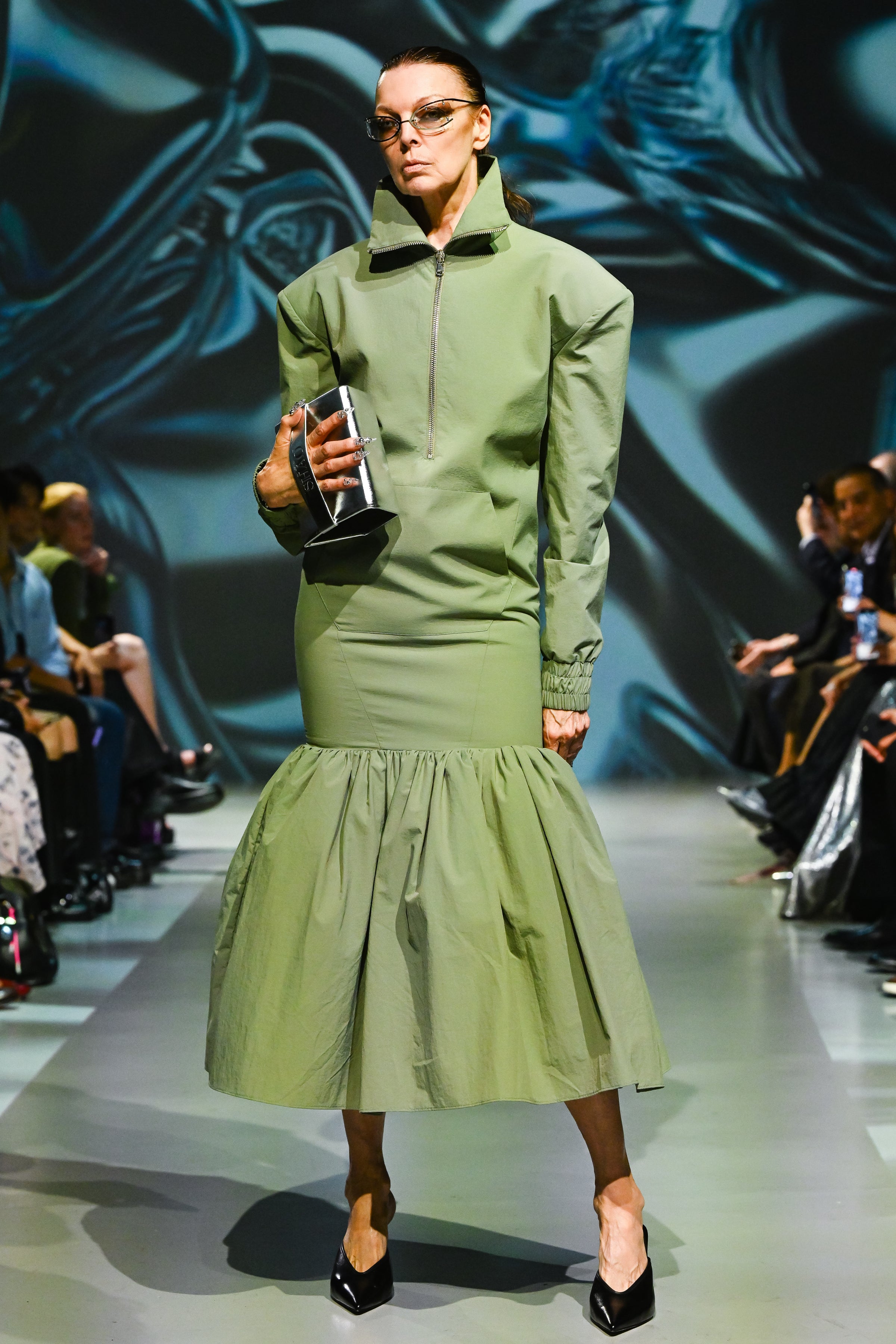 Model walks runway wearing an olive-green structured dress with exaggerated padded shoulders, a high collar, front zipper, and a gathered flared hem, paired with black heels and the SHAO X Dagne Dover Silver Metallic Eyewear Clutch Bag, exemplifying SHAO New York luxury designer fashion and sculptural silhouette.