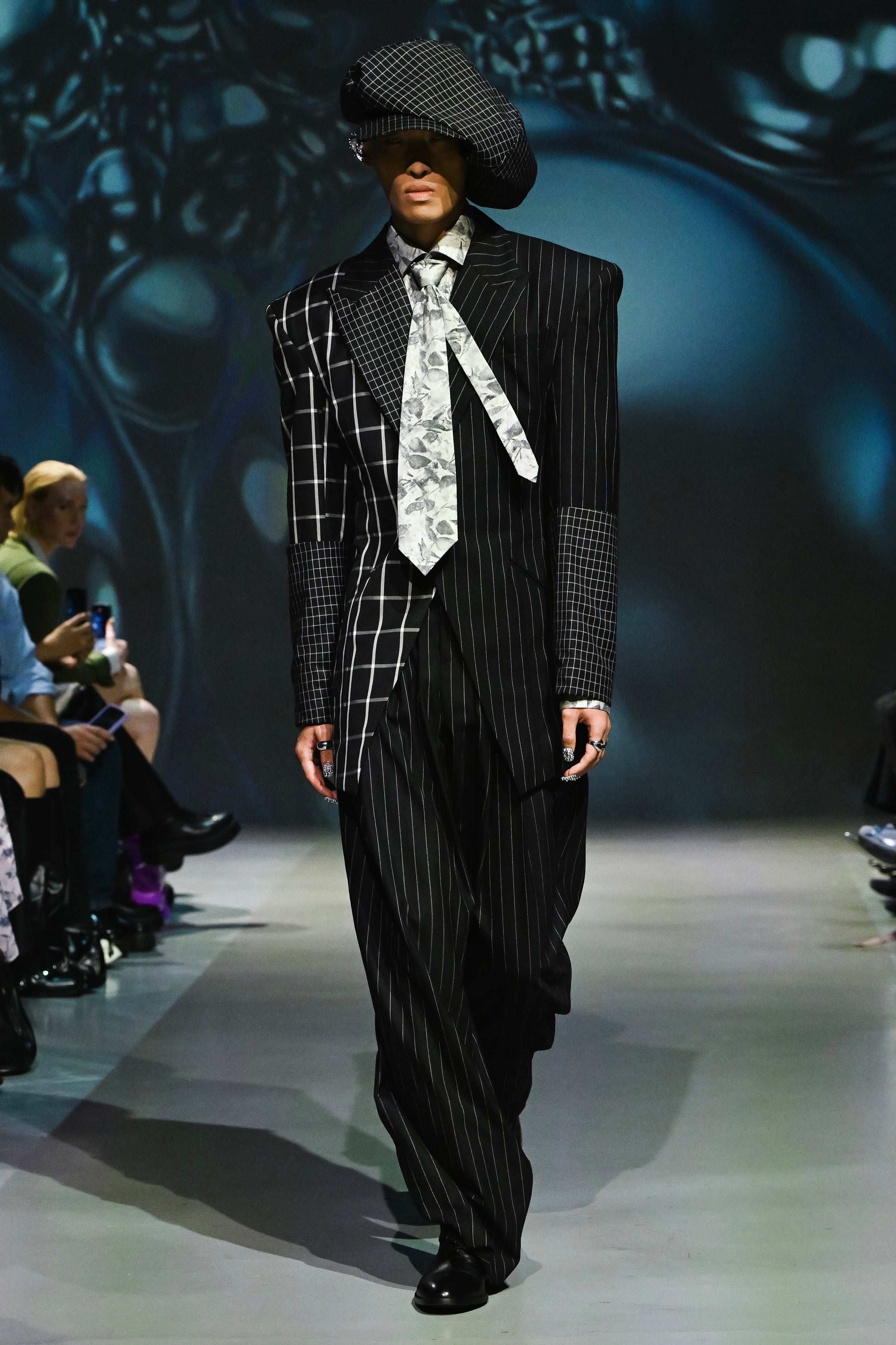 Model wearing a pinstripe suit and hat on a runway with a dark, abstract background.
