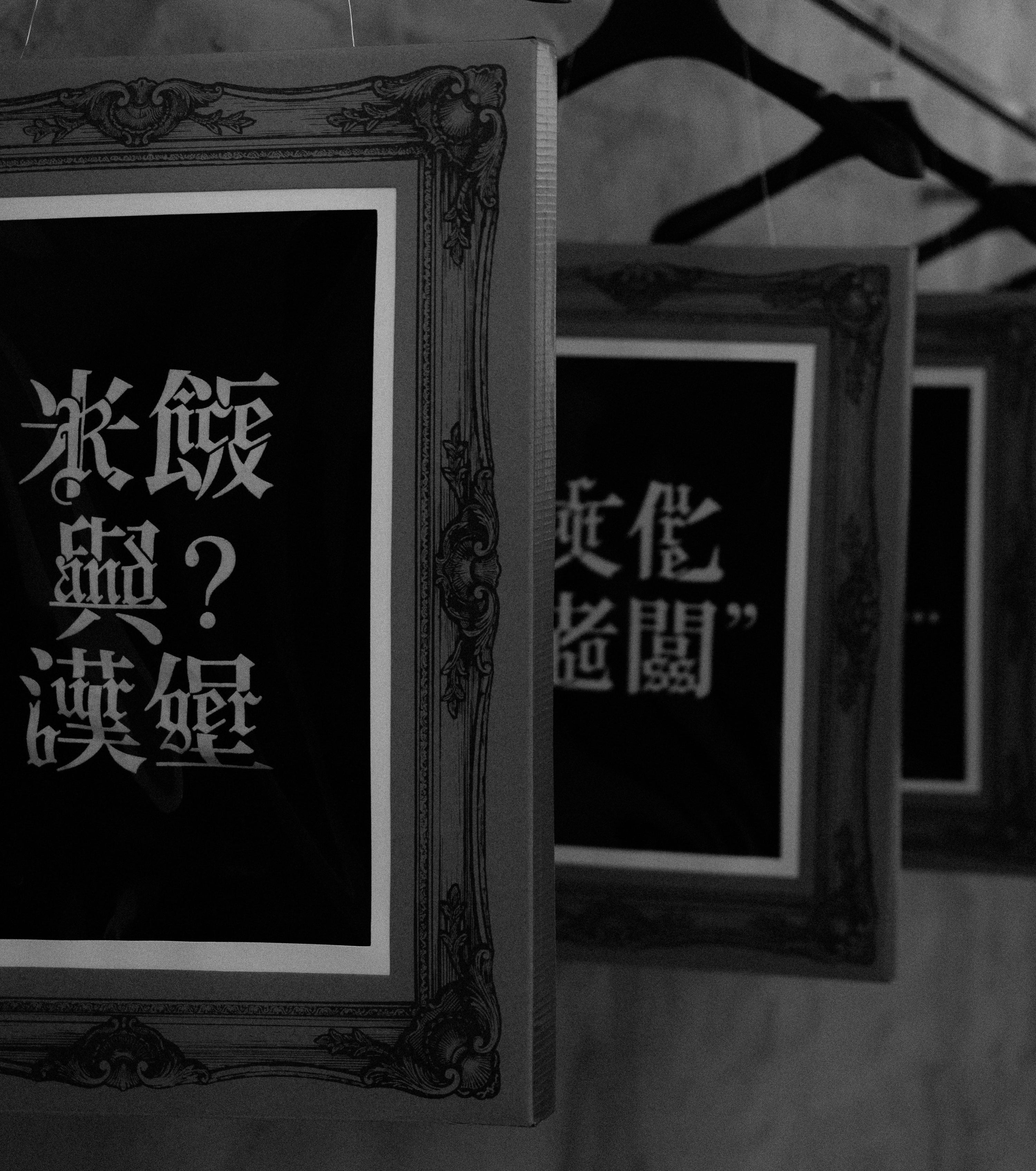Three framed prints featuring stylized Asian characters and English text on black backgrounds, arranged in a row on a textured wall with soft lighting.