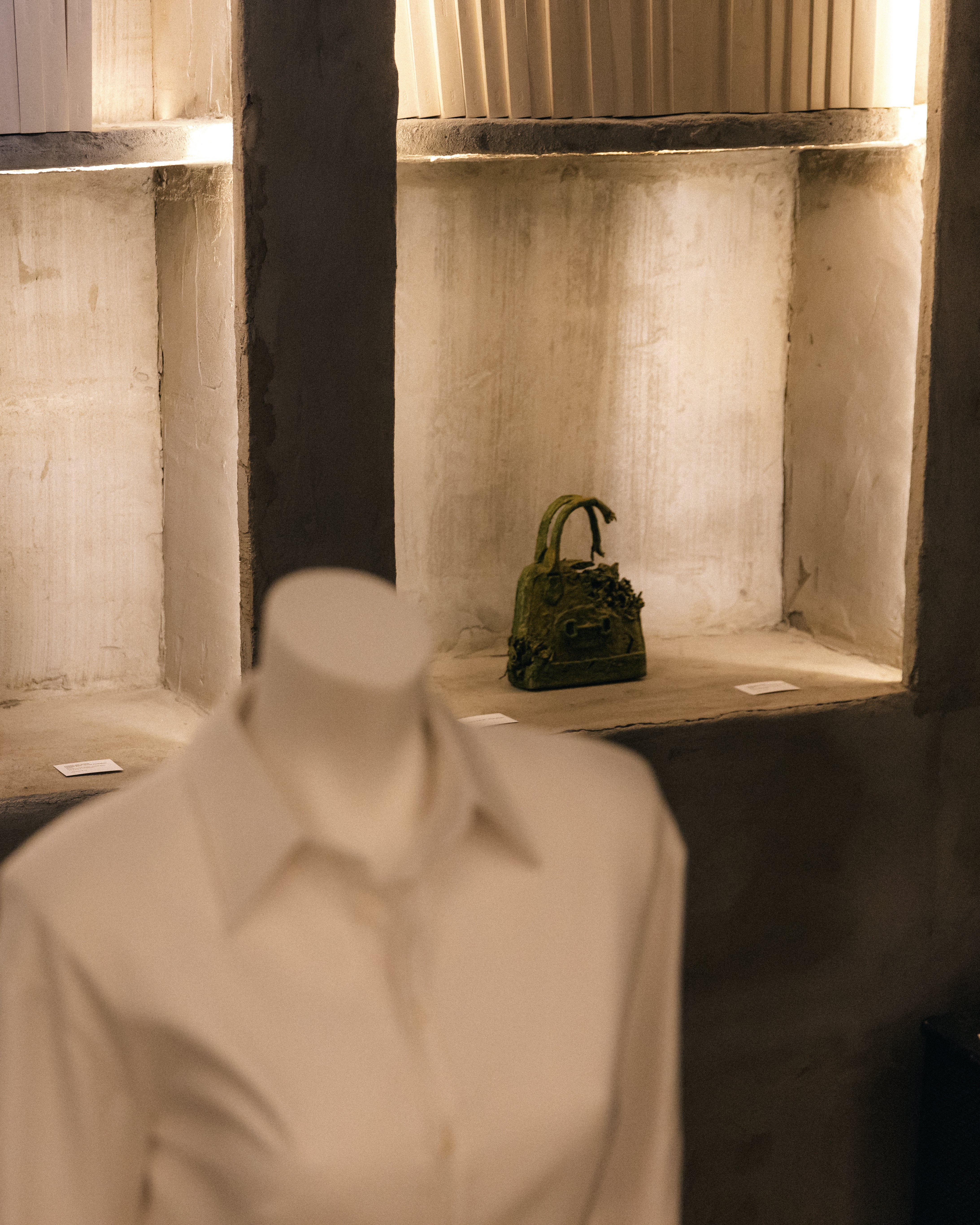 Softly lit concrete niches displaying an intricately textured green handbag with a blurred mannequin dressed in a cream shirt in the foreground, evoking an artistic boutique atmosphere.