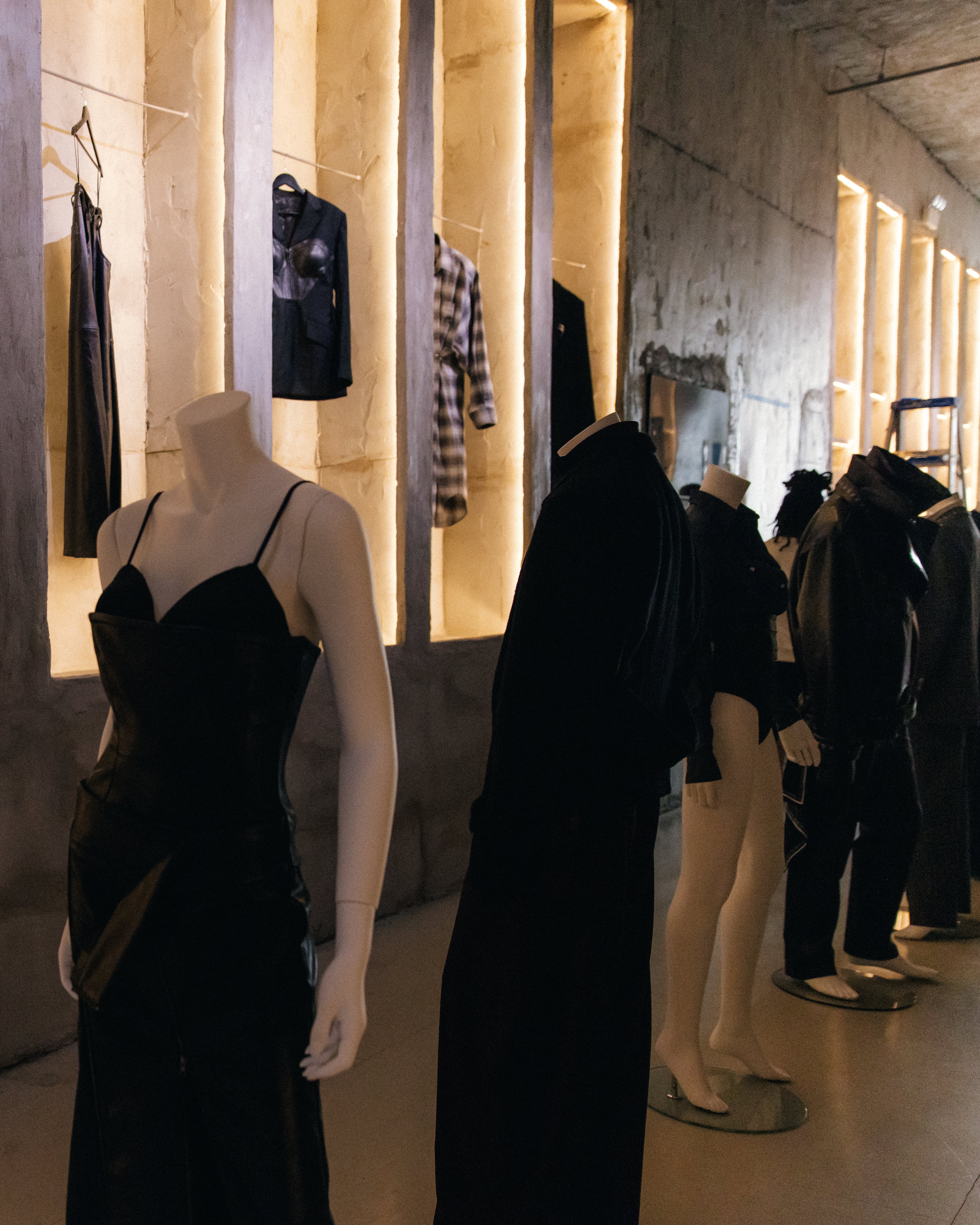 A row of mannequins dressed in dark, tailored garments including a sleeveless dress and structured jackets, positioned in a dimly lit space with vertical illuminated wall niches displaying additional clothing pieces.