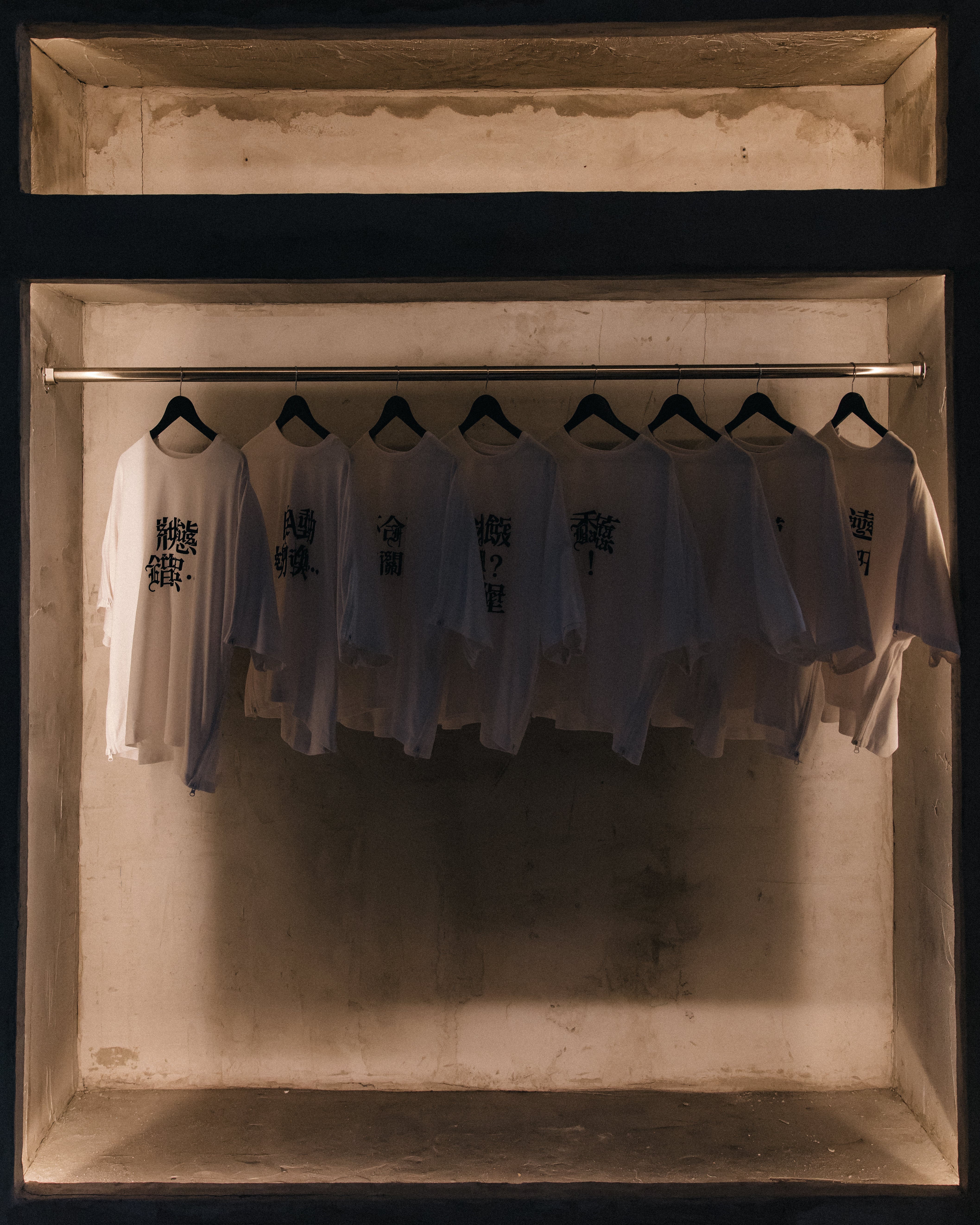 Row of white long-sleeve shirts with black stylized Asian characters printed on the front, hanging evenly spaced on black hangers from a horizontal rod inside a dimly lit, rustic recessed display space.