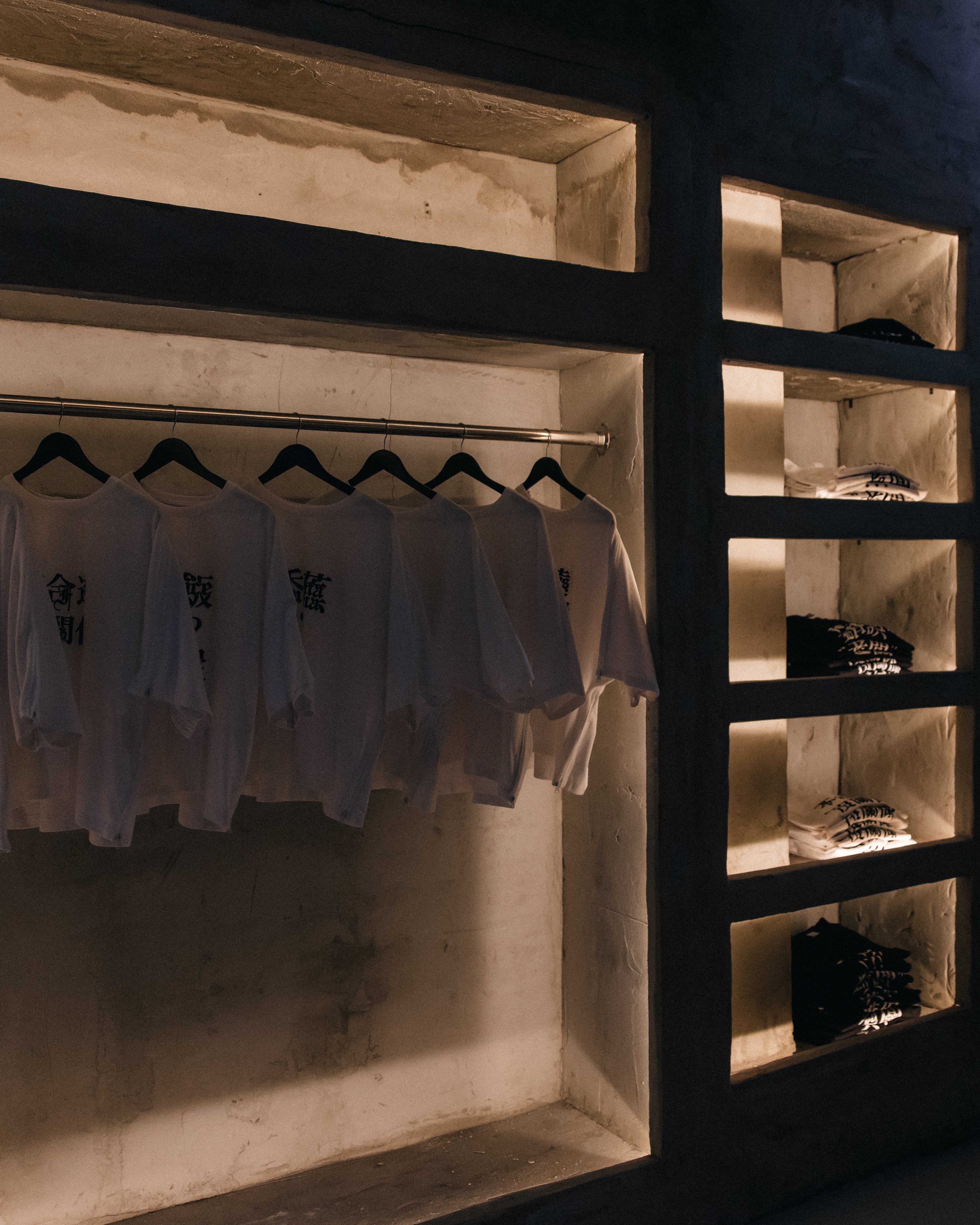Dimly lit display of white and black garments arranged on hangers and shelves recessed into a textured concrete wall, highlighting minimalist industrial design.