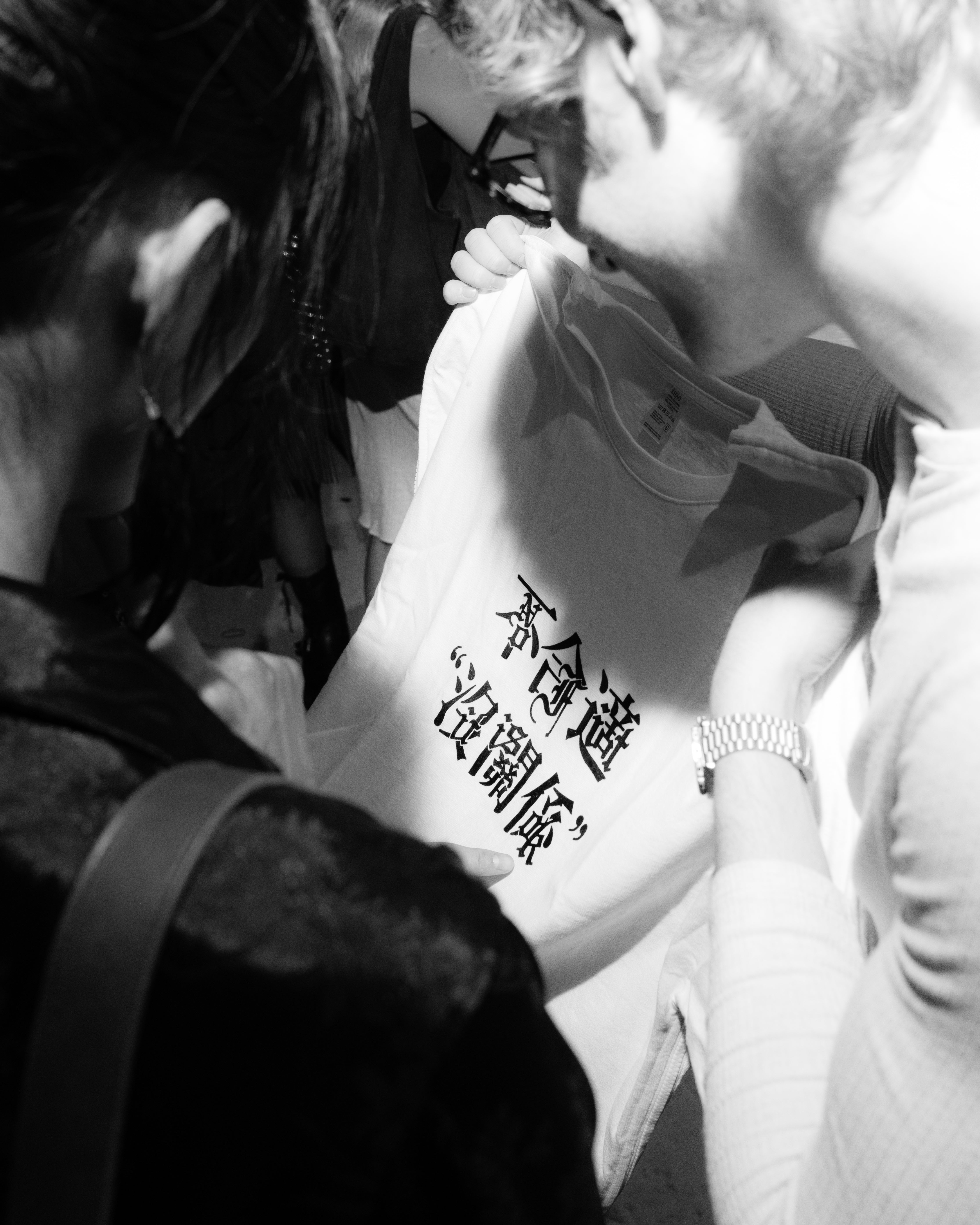 Two people closely examine a white garment with bold black Asian characters printed on the fabric, captured in soft black-and-white lighting.