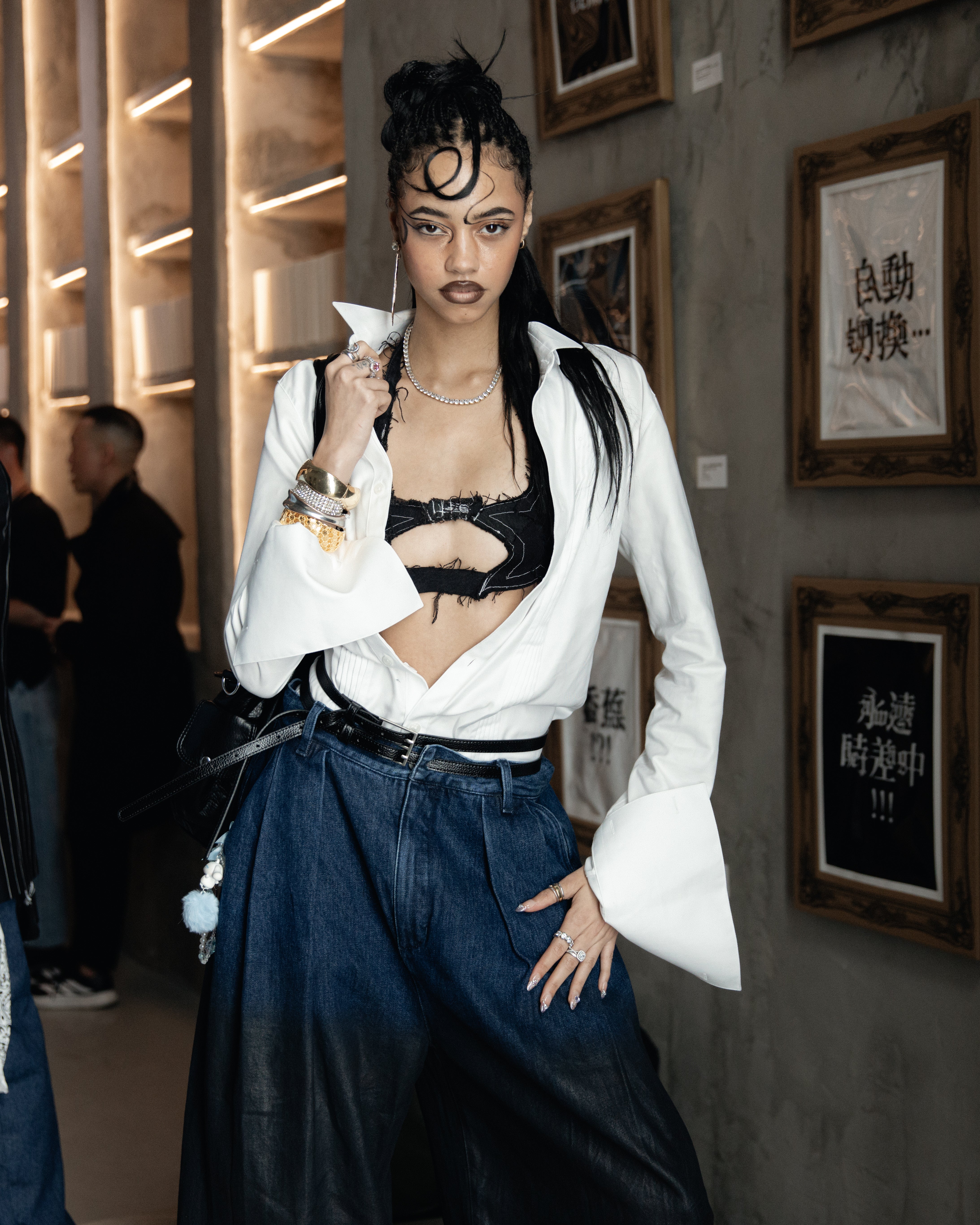 Tall model with styled hair wearing a white shirt with exaggerated cuffs over a black cutout top and high-waisted wide-leg denim pants, posing indoors near framed calligraphy artwork.
