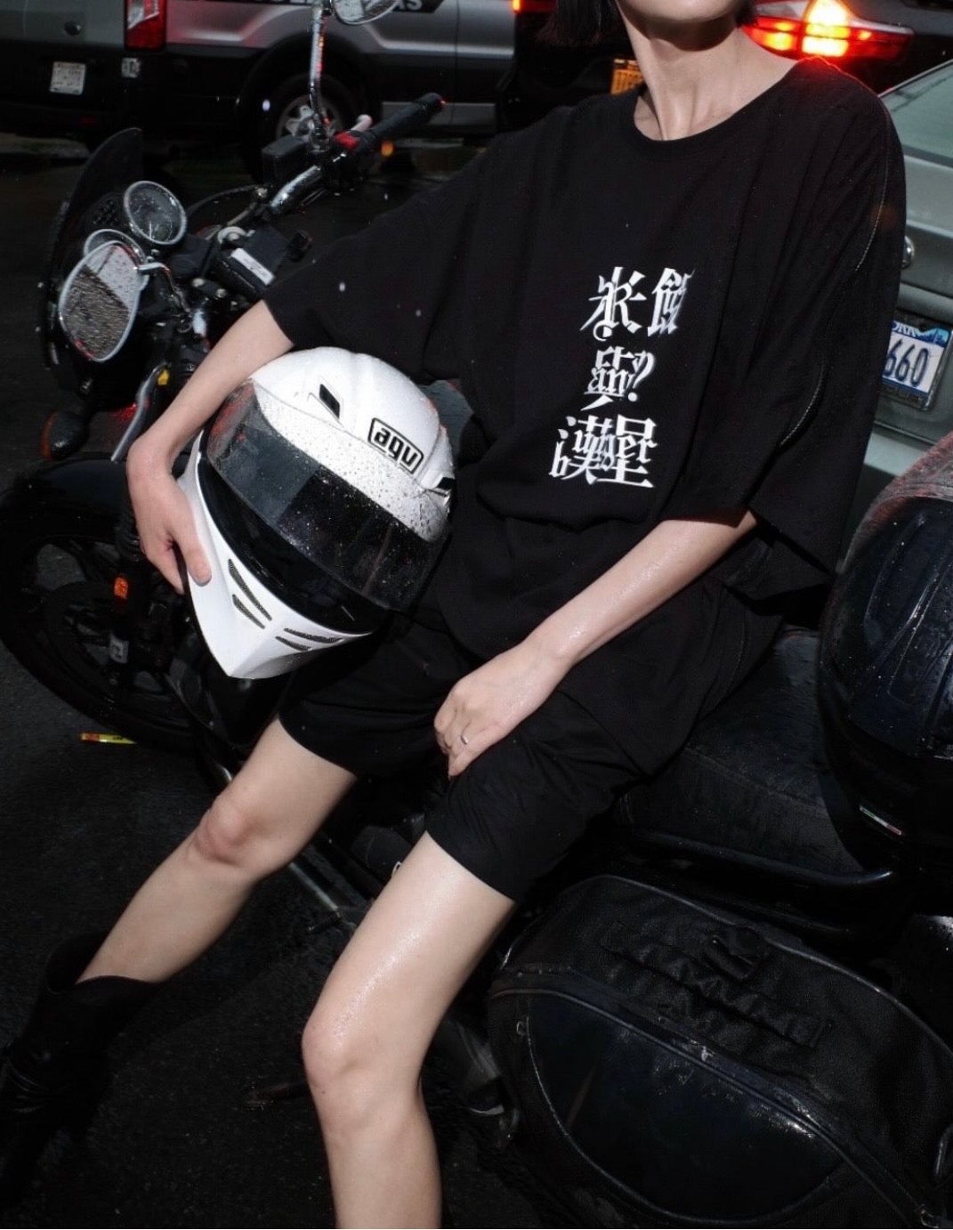 Person wearing a loose black shirt with white Asian characters printed on the chest, black shorts, and holding a white motorcycle helmet while sitting on a black motorbike on a wet street at night.
