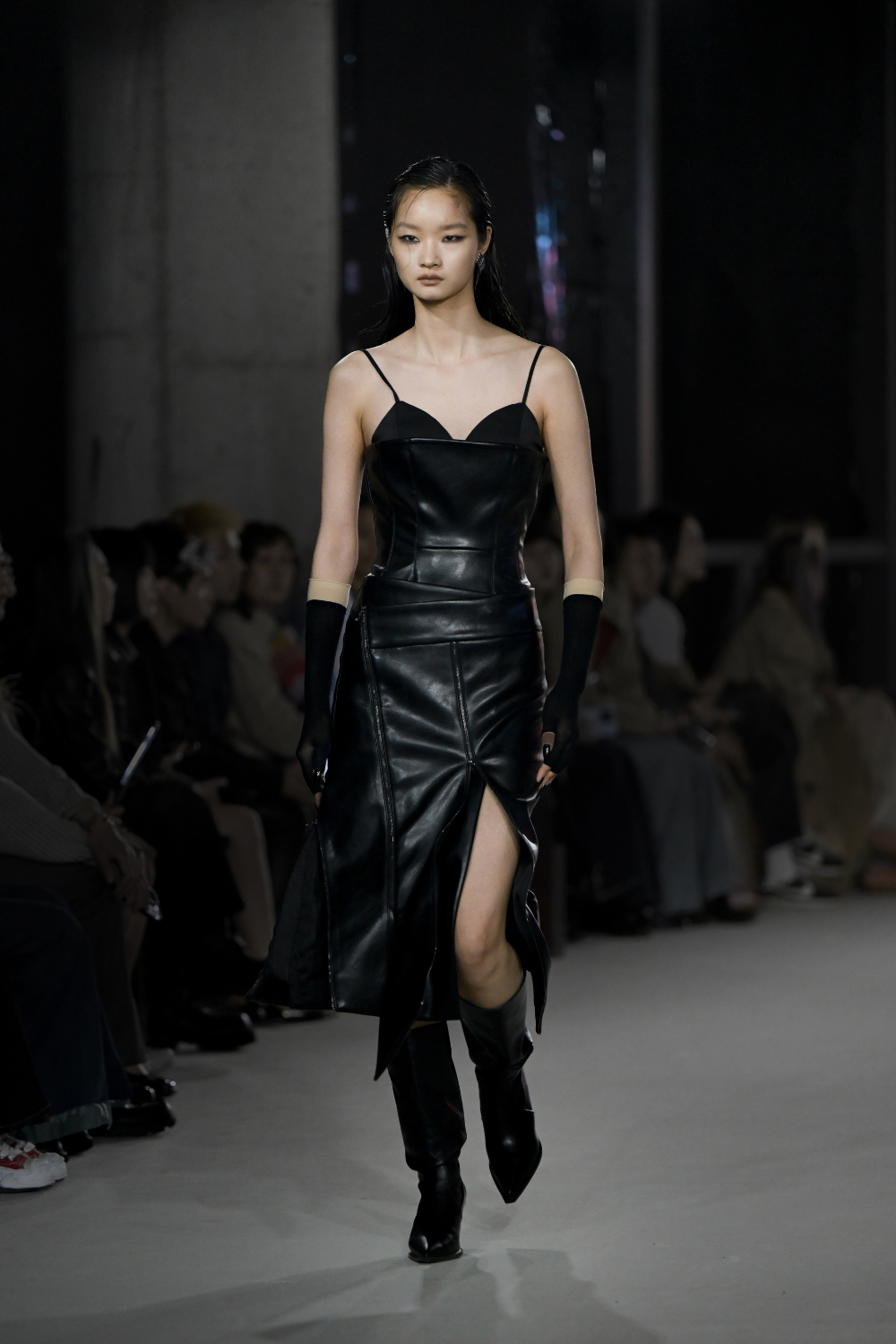 Model walking runway wearing the Vegan Leather Biker Dress featuring thin straps, a fitted structured bodice, a front thigh-high slit, paired with long fingerless gloves and black boots, showcasing SHAO New York bespoke tailoring and a sculptural silhouette.