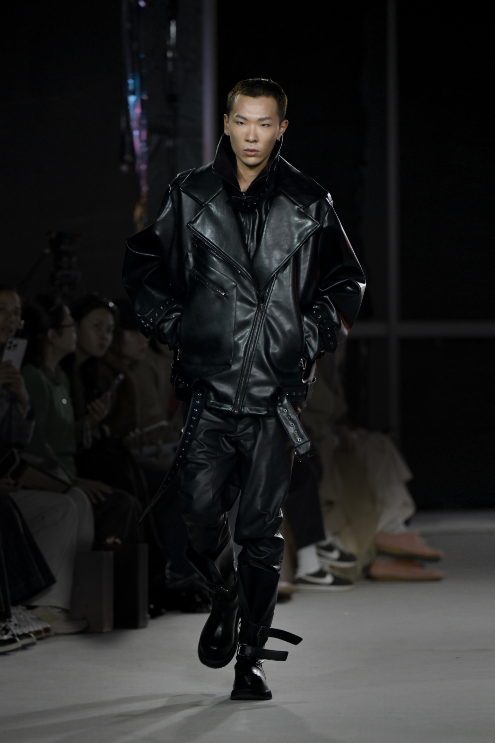 Model walks runway wearing the Vegan Leather Oversized Biker Coat in black with bold sculptural lapels, asymmetrical zipper, and adjustable buckle straps, paired with matching pants, showcasing SHAO New York luxury designer fashion and expert New York designer tailoring.