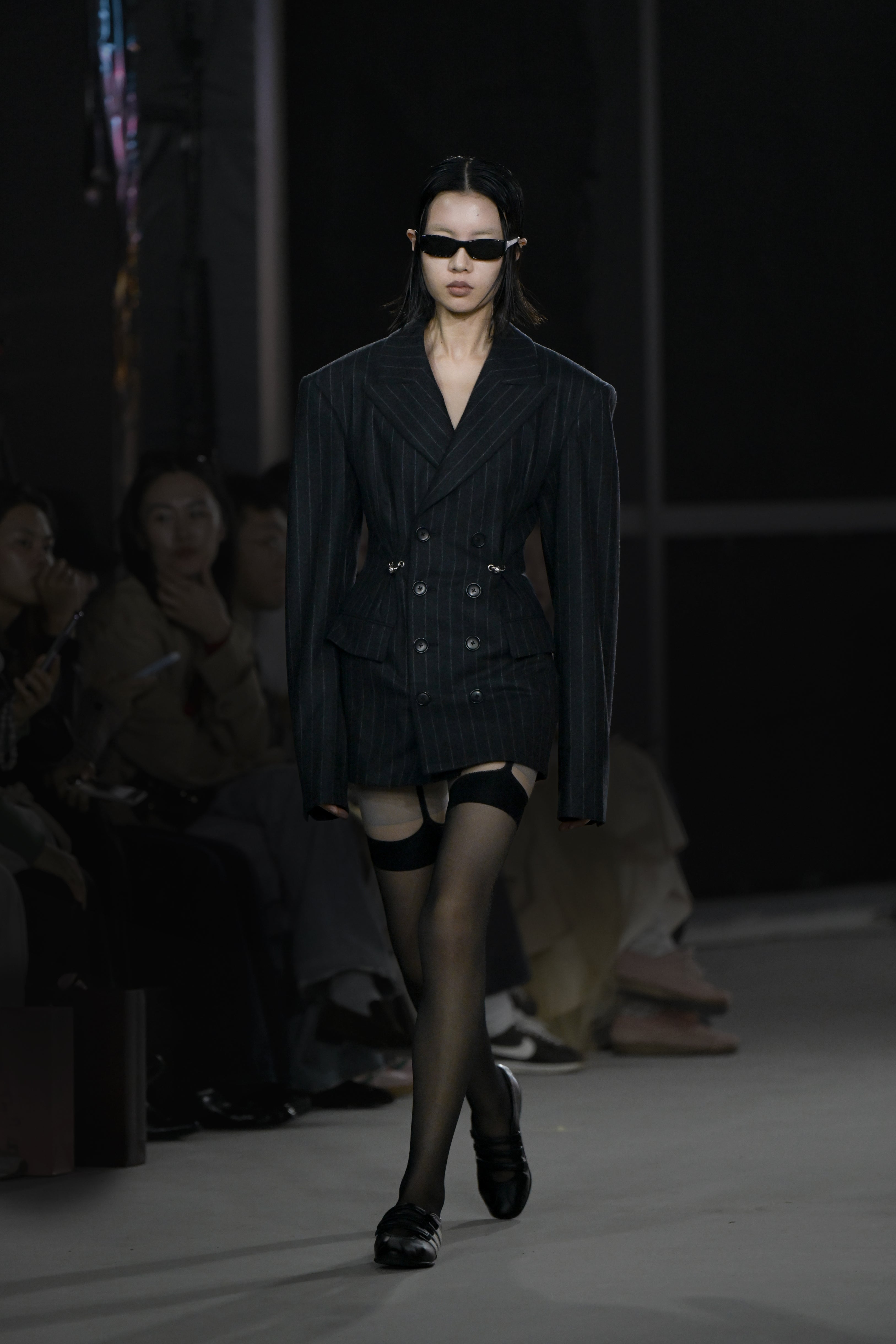 A model walks the runway wearing an oversized pinstripe blazer with exaggerated shoulders and double-breasted buttons, paired with thigh-high sheer stockings, black loafers, and small black sunglasses.