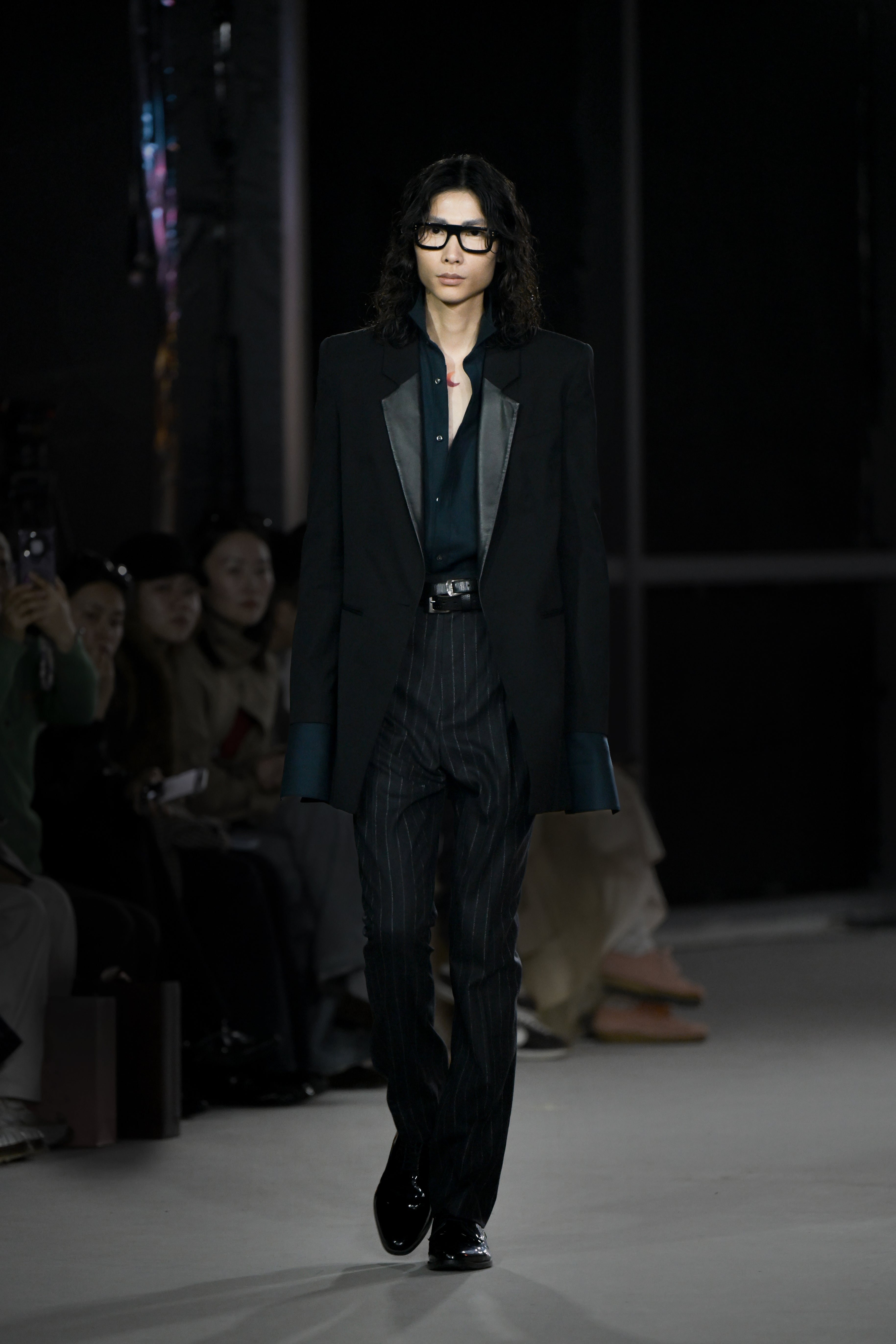 A model walks the runway wearing a tailored black blazer with contrasting lapels over a dark shirt and pinstriped trousers, styled with black glasses and polished shoes, presenting a refined luxury silhouette.