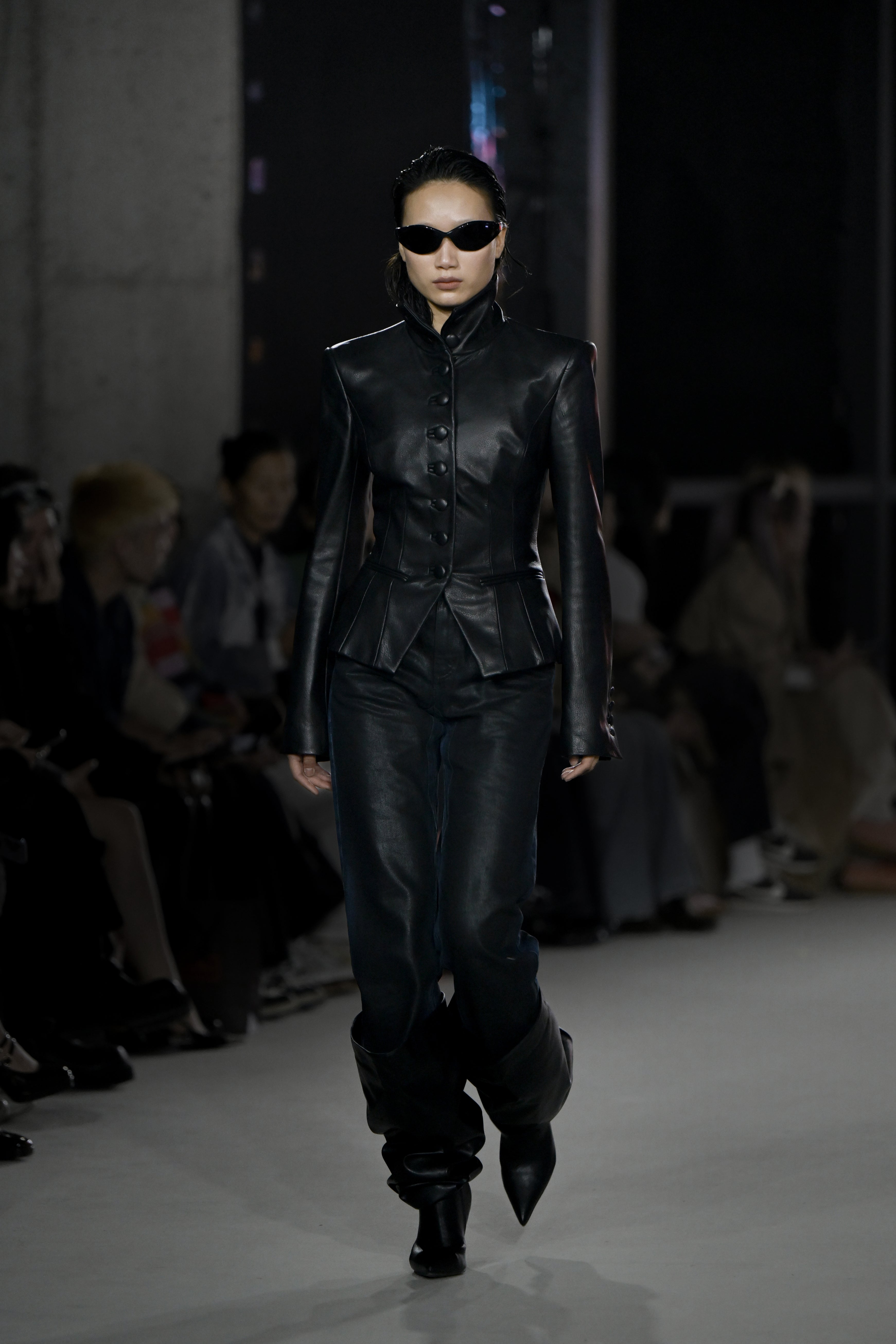 A model walks the runway wearing a structured black leather jacket with a high collar and buttoned front, paired with matching black leather trousers and pointed boots, styled with narrow black sunglasses for a sleek sculptural silhouette.