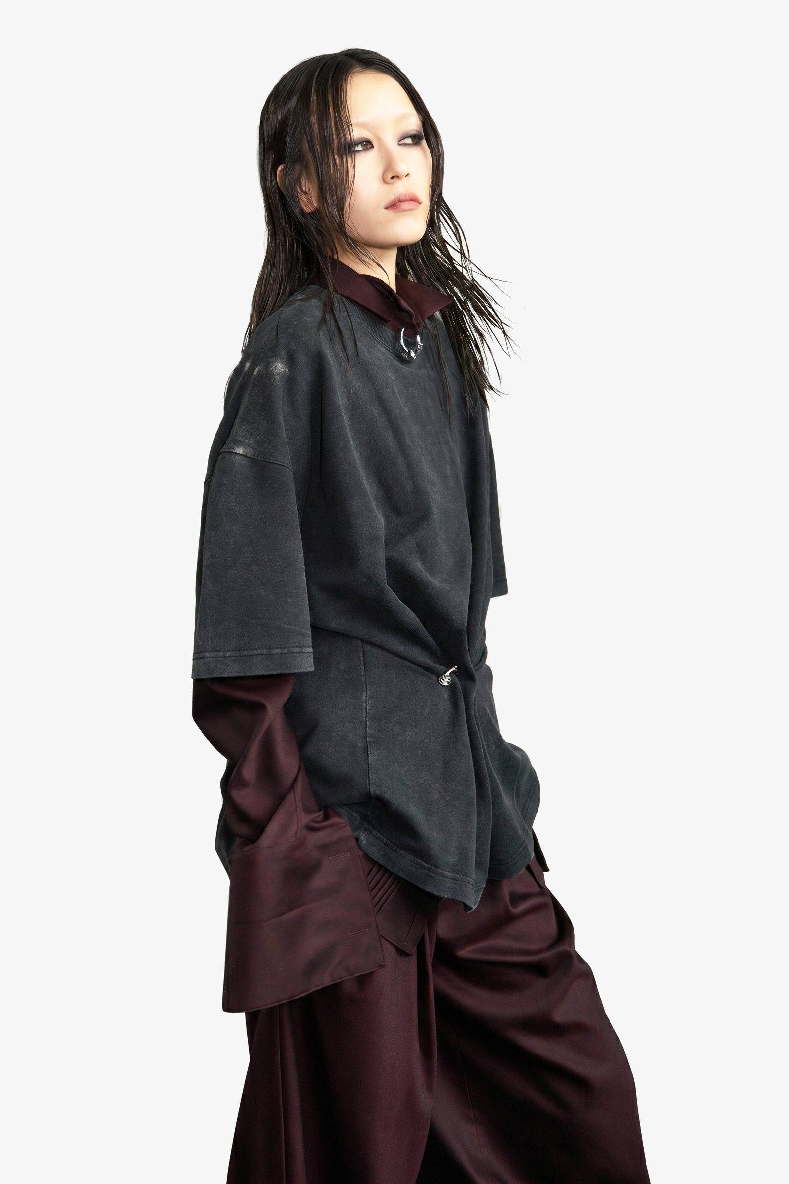 Model wearing layered oversized black washed shirt over burgundy satin long-sleeve top and wide-leg trousers, styled with a minimal Piercing accessory, embodying avant-garde luxury designer fashion.
