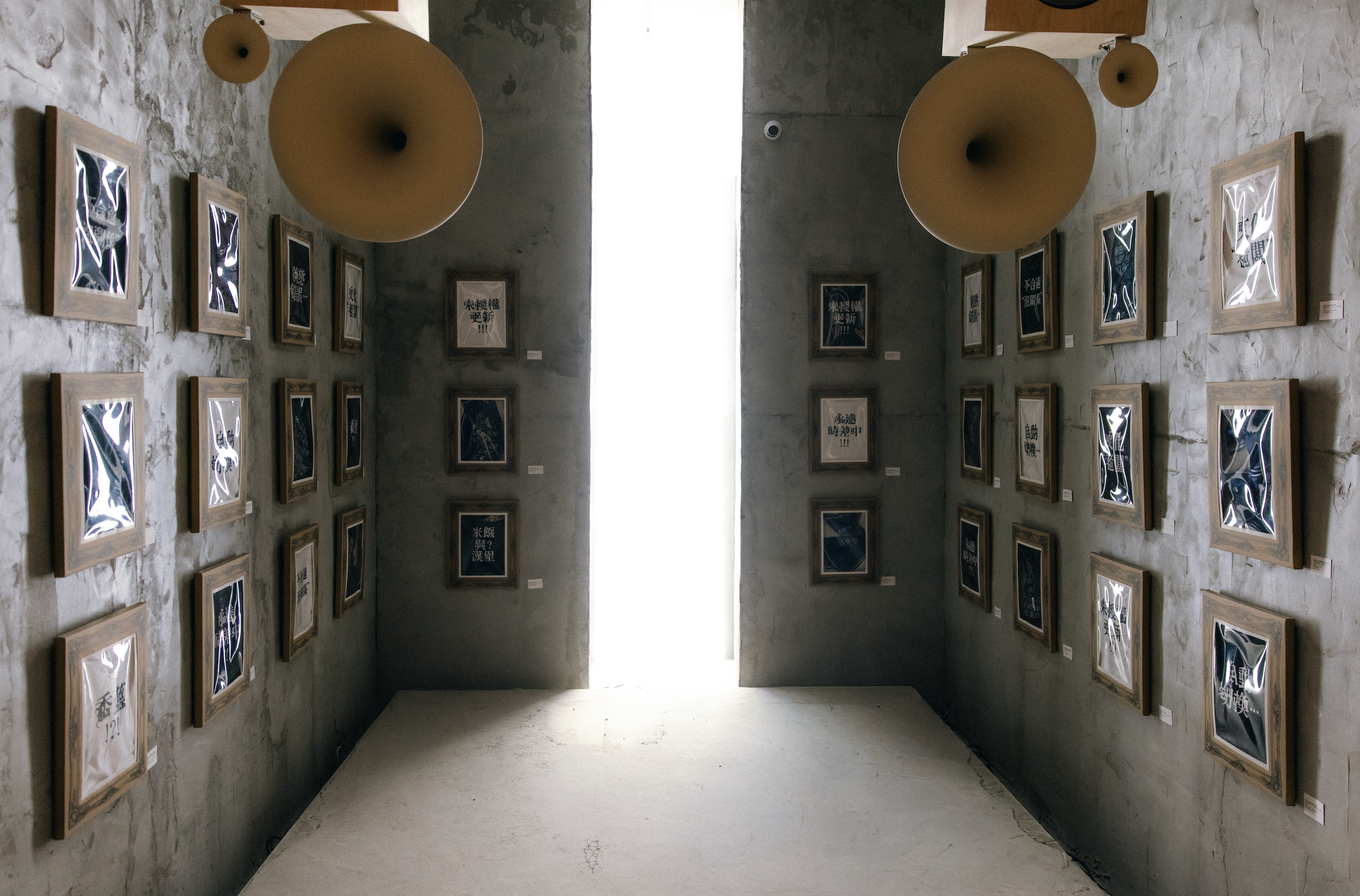 Minimalist art gallery corridor with framed abstract prints lining textured concrete walls, illuminated by soft natural light from a tall central window and overhead circular speakers.