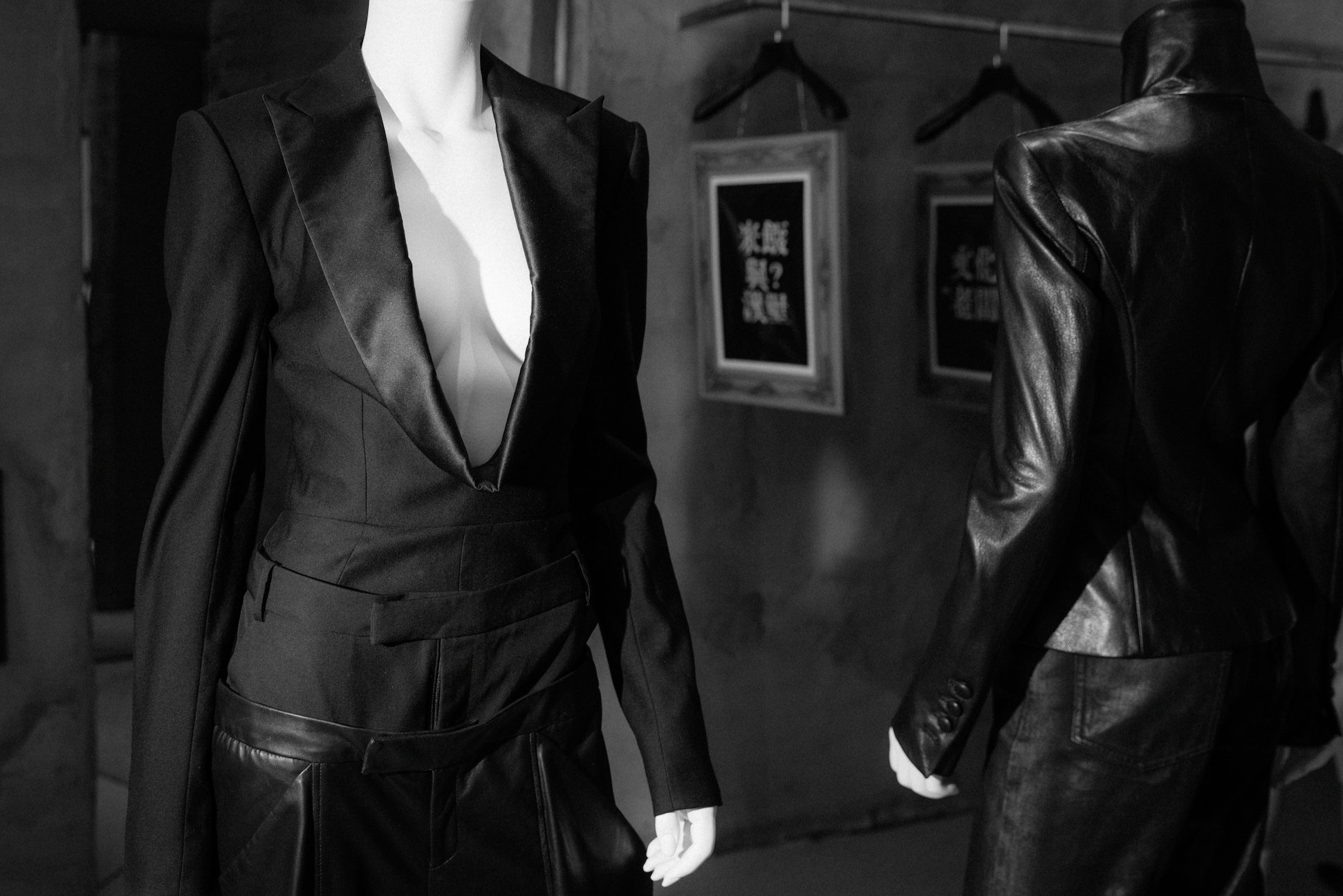 Two mannequins dressed in tailored black outfits with structured silhouettes, displayed in a dimly lit space featuring framed calligraphy art on textured walls.
