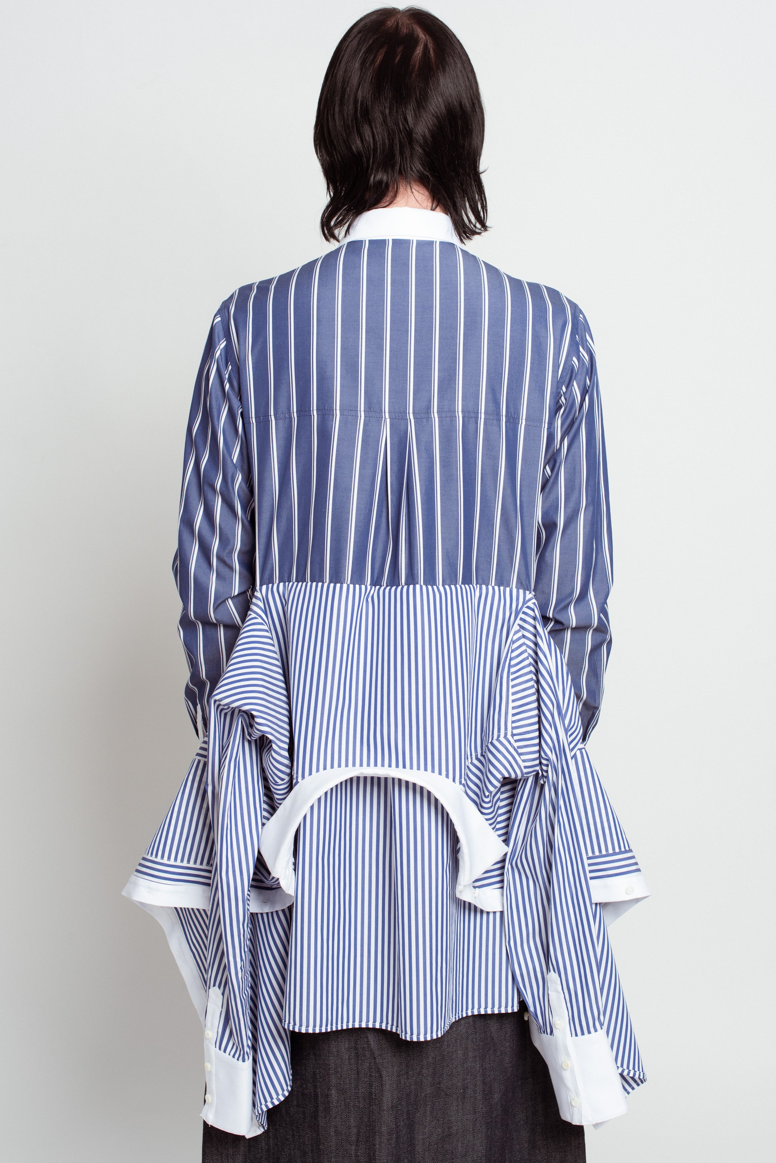 Back view of Blue Pinstripe Layered Shirt – Attached Back Construction featuring contrasting vertical stripes, draped layered fabric detail, crisp white collar and cuffs, showcasing SHAO New York bespoke tailoring and sculptural silhouette.