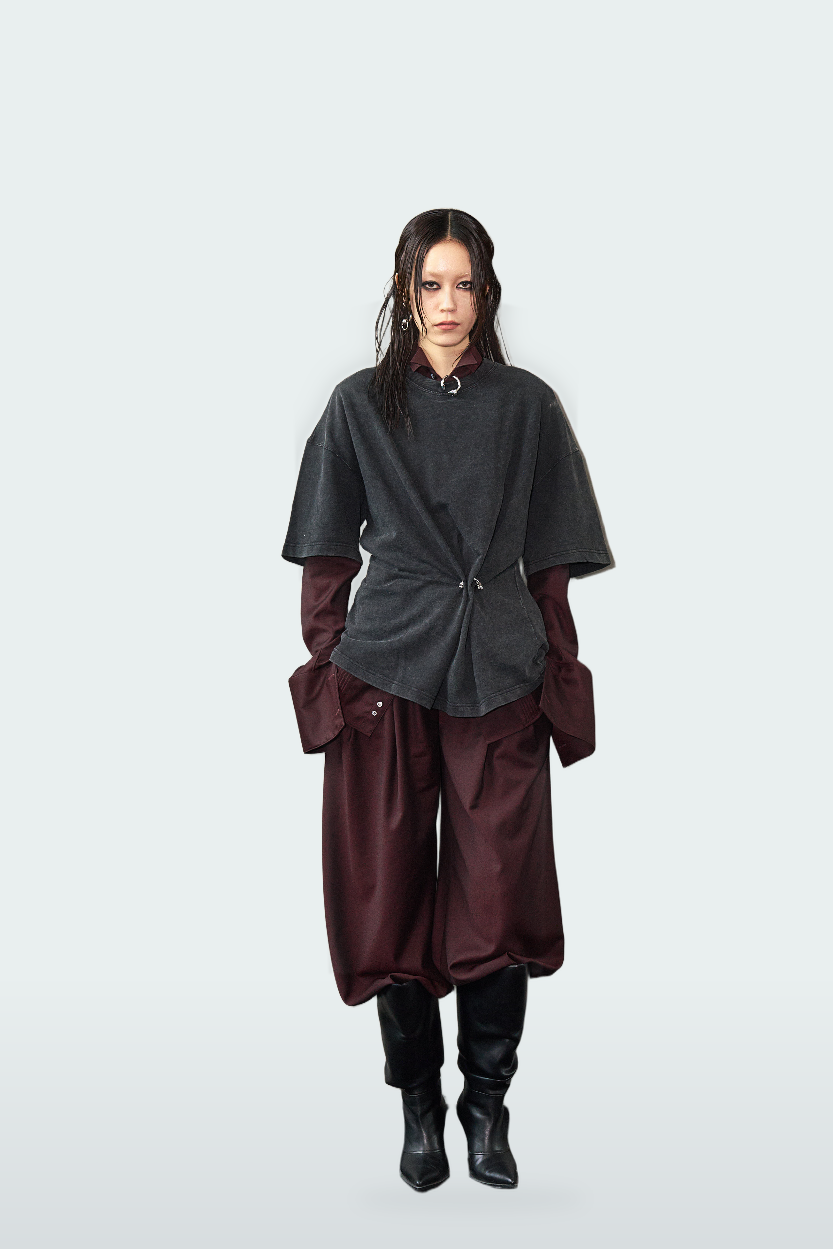 A model stands against a light gray studio background wearing a dark charcoal gray wrap-style top with dark red sleeves and matching dark red wide-leg pants, paired with black boots.