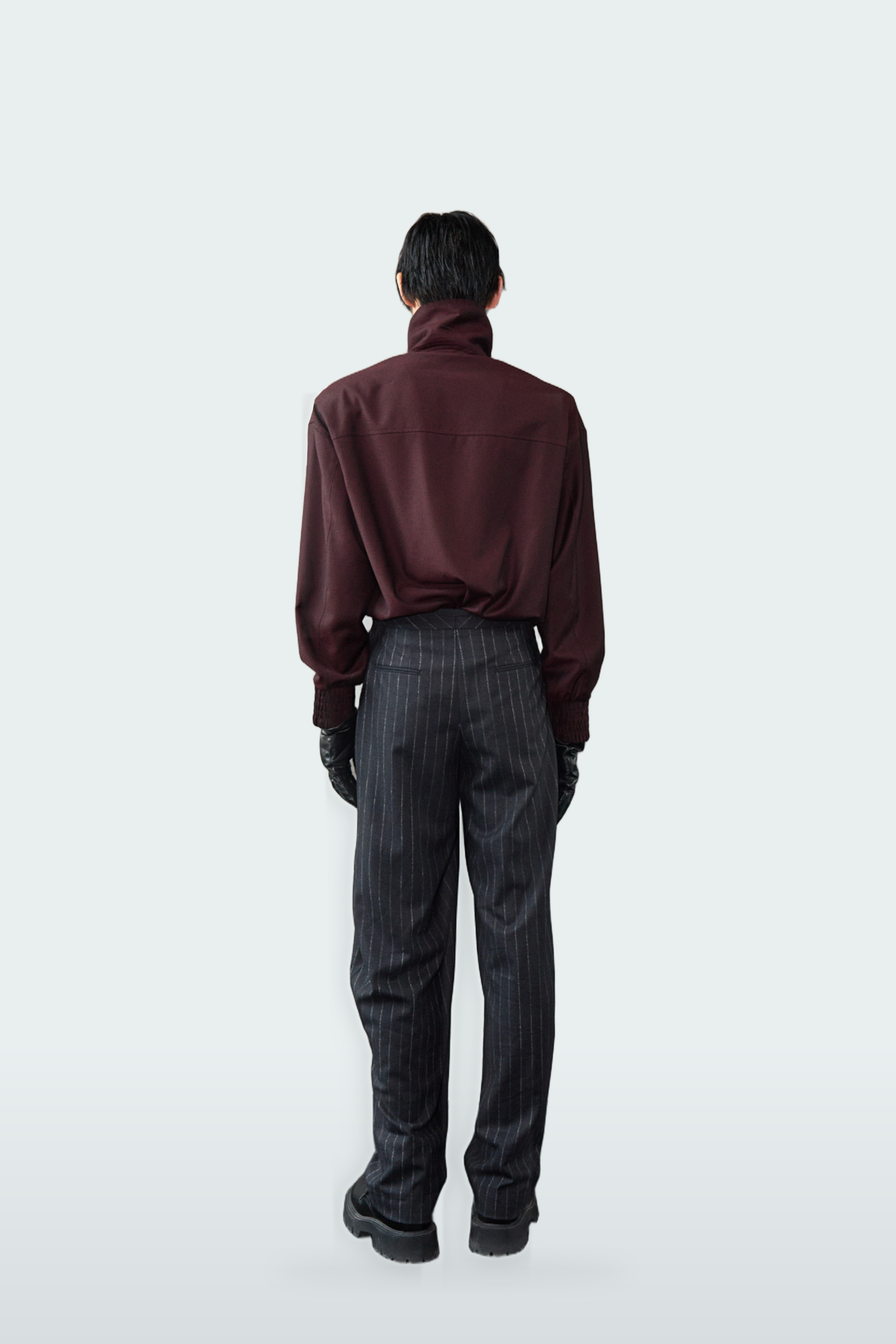 A model viewed from behind wearing a dark burgundy turtleneck shirt, dark gray pinstriped trousers, and black gloves, set against a solid light gray background.