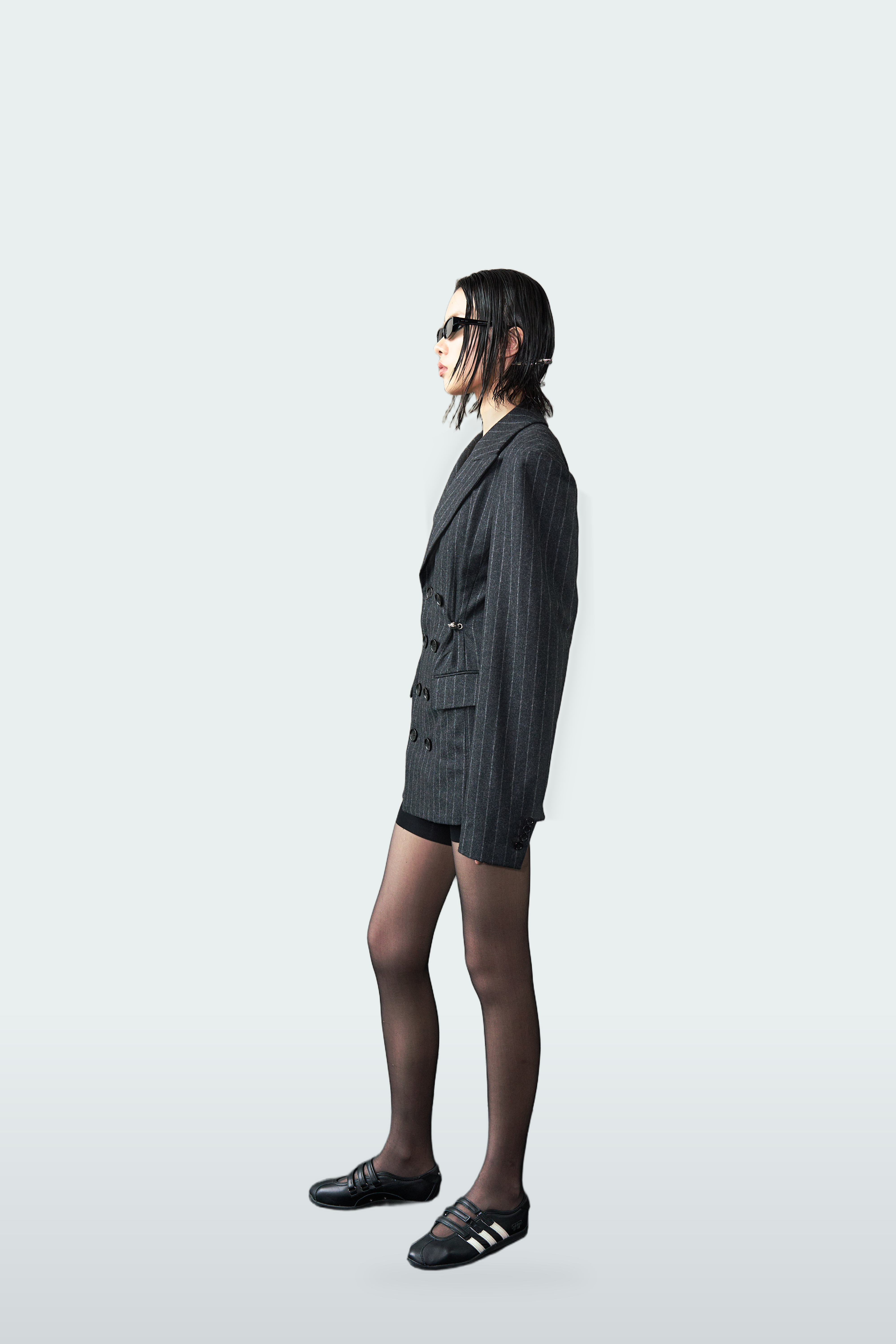 A model is shown in profile wearing a dark gray pinstripe double-breasted blazer, black shorts, sheer black tights, and black slide sandals with white stripes, set against a solid light gray background.