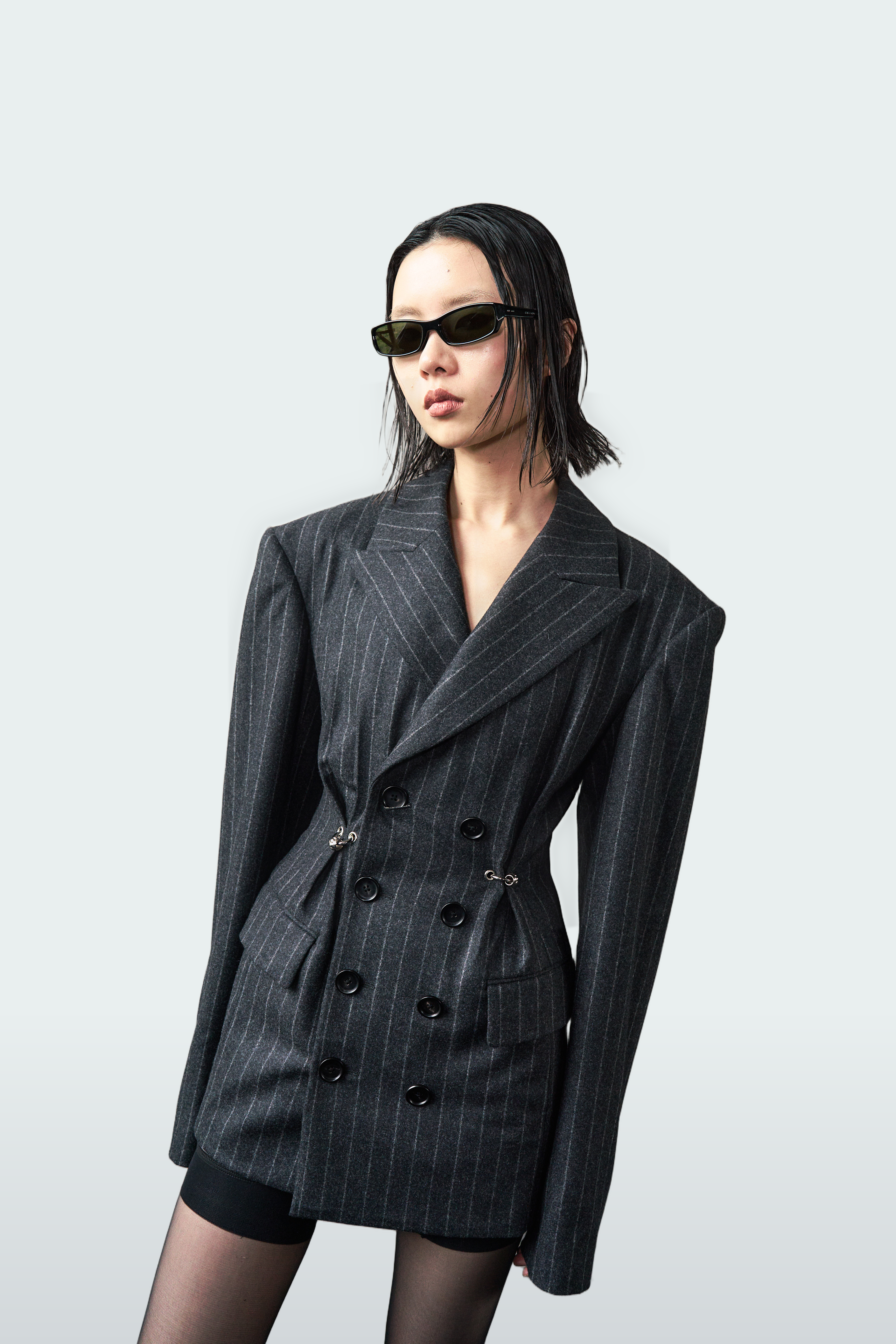A model wearing a dark gray double-breasted pinstripe blazer with matching mini skirt, black sunglasses, and sheer black tights, posed against a light gray studio background.