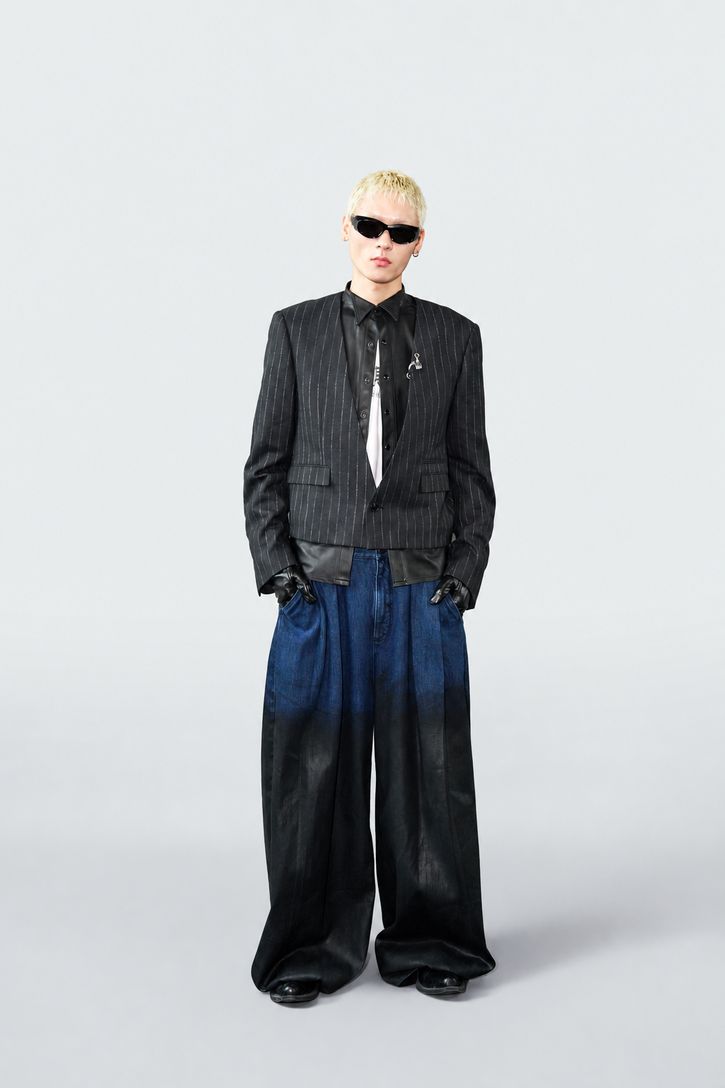 A model with short blonde hair and sunglasses stands against a light gray studio background, wearing a black pinstripe blazer, black shirt and tie, wide-leg black pants with a blue-to-black gradient upper section, and black gloves.
