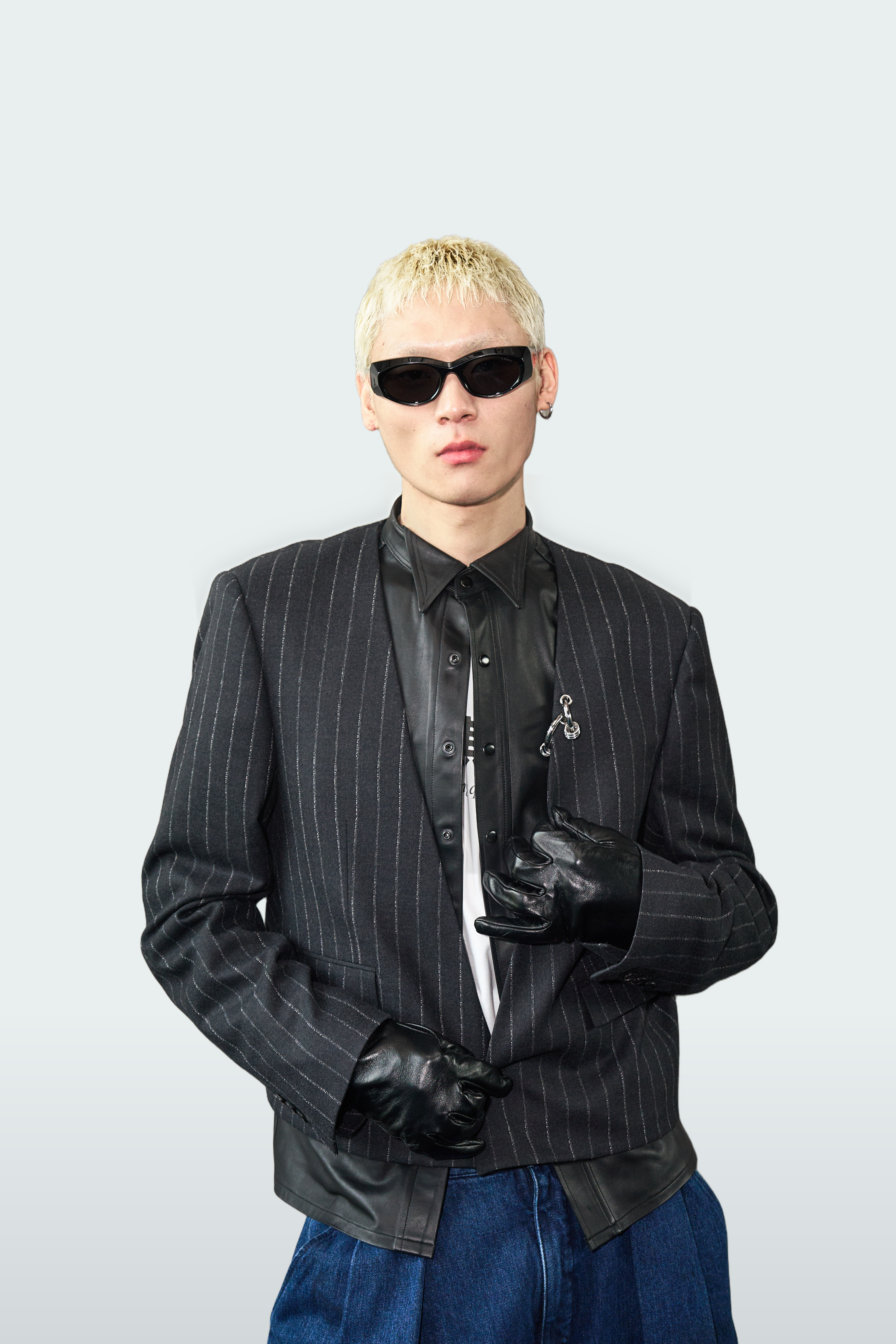 A model with short blonde hair wearing a black pinstripe blazer, black leather shirt, black tie, black gloves, and sunglasses, paired with blue jeans against a light gray background.