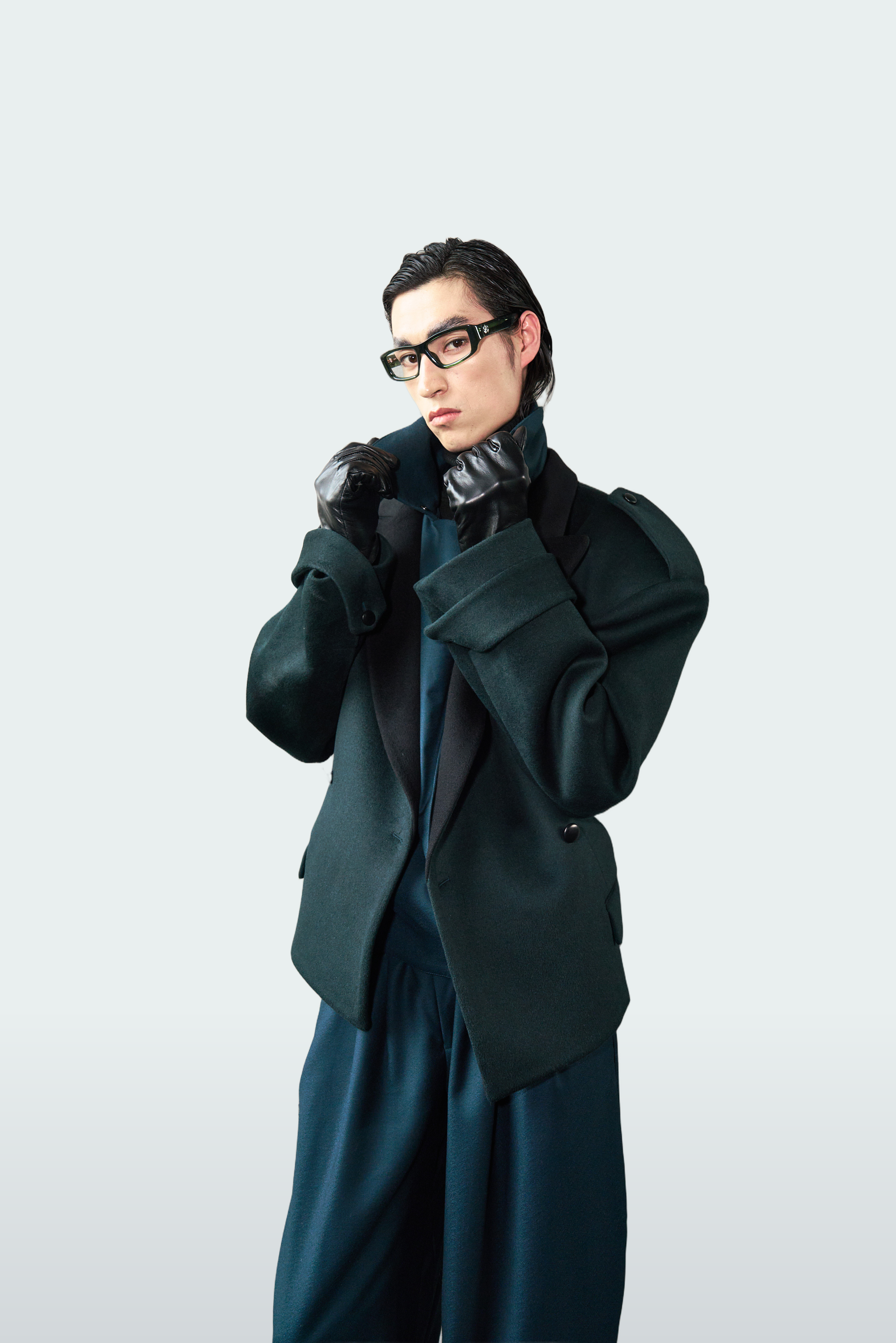 A model wearing a dark green oversized tailored jacket with black lapels, matching dark green wide-leg trousers, black gloves, and black-rimmed glasses, posed against a light gray studio background.