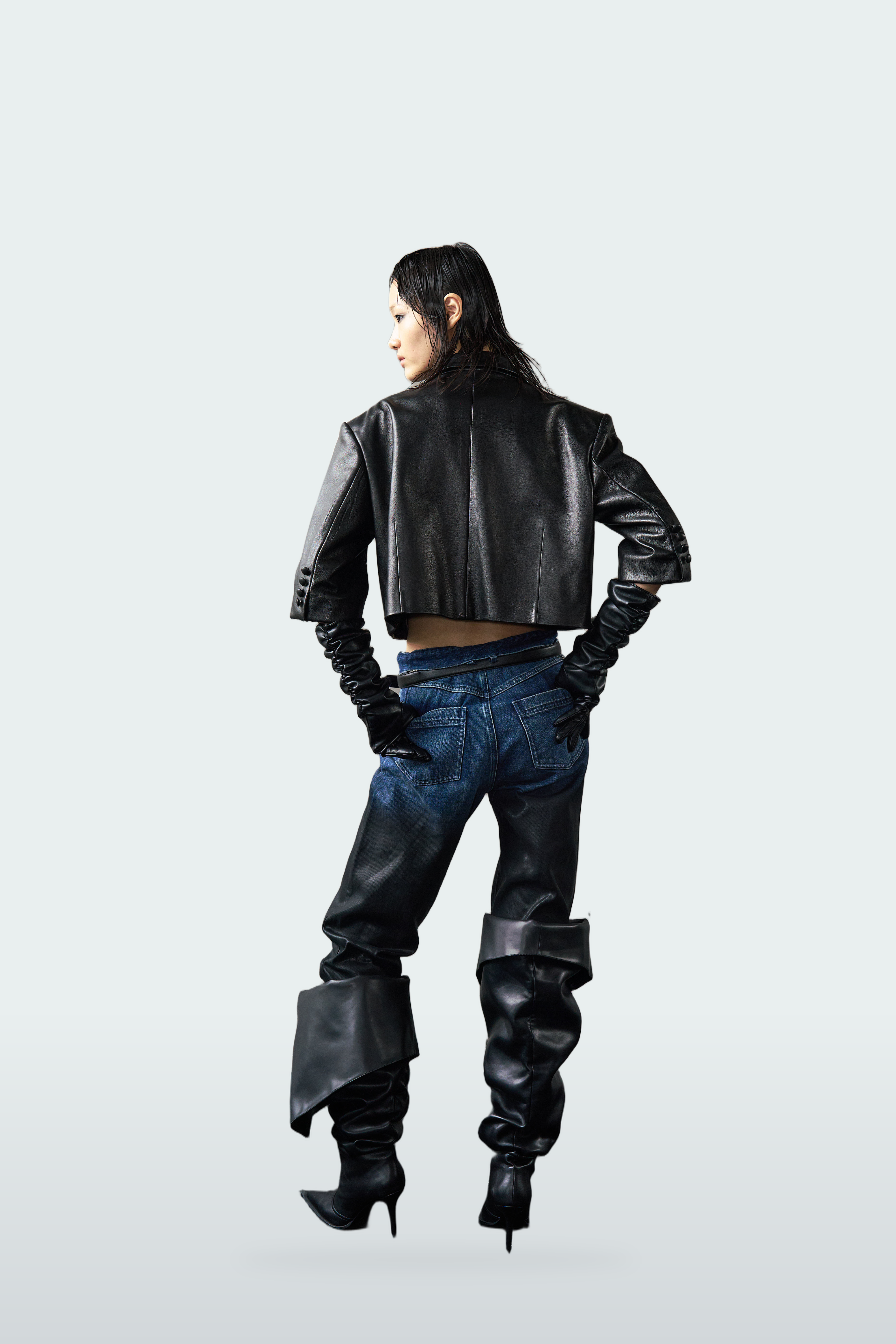 A model is shown from behind wearing a black cropped leather jacket, dark blue jeans, black leather pants with unique folded leg details, and black high-heeled boots against a light gray studio background.