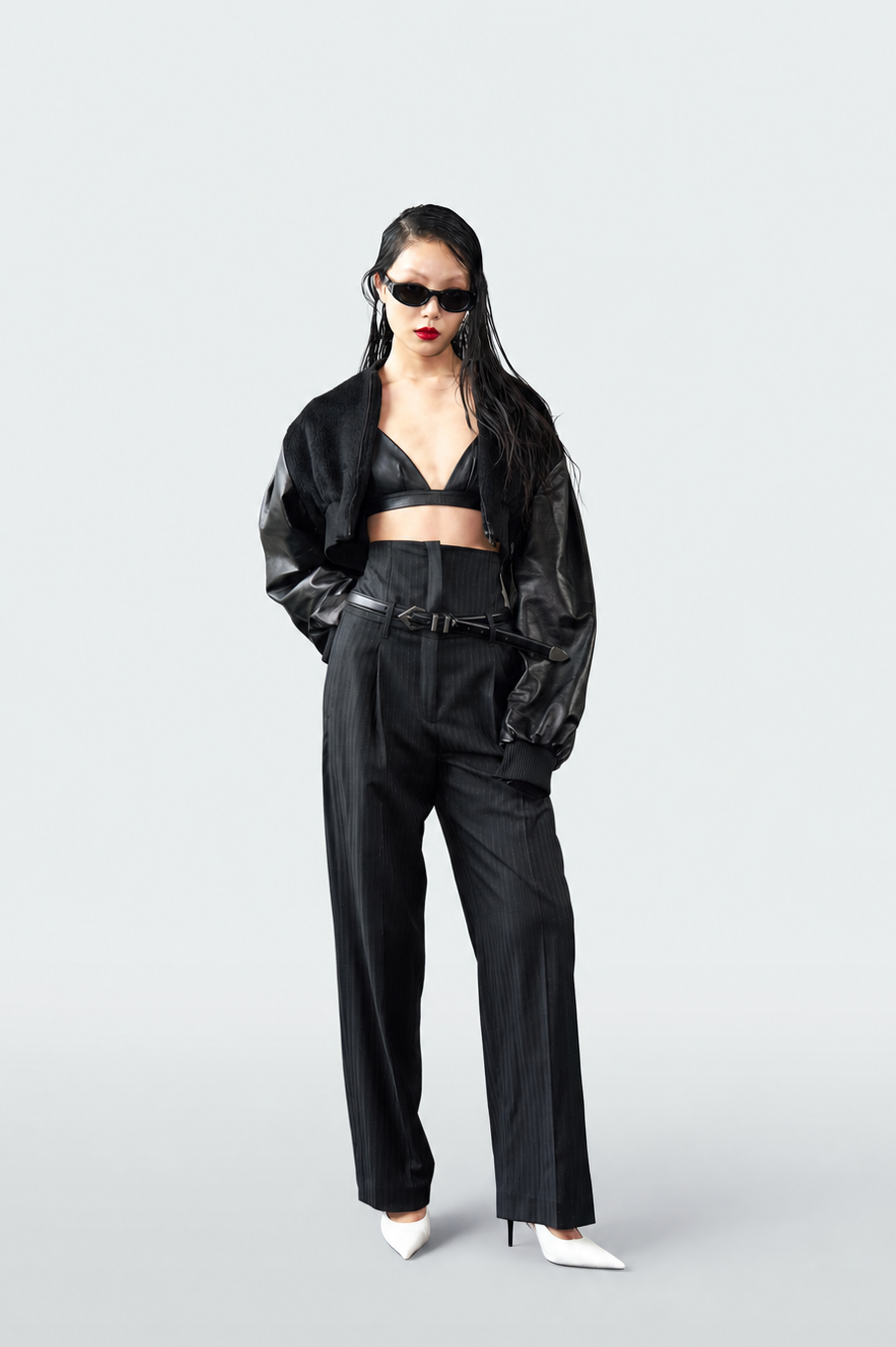 A model stands against a light gray studio background wearing a black cropped jacket over a black bralette, black high-waisted wide-leg trousers with a matching belt, and white pointed-toe heels.