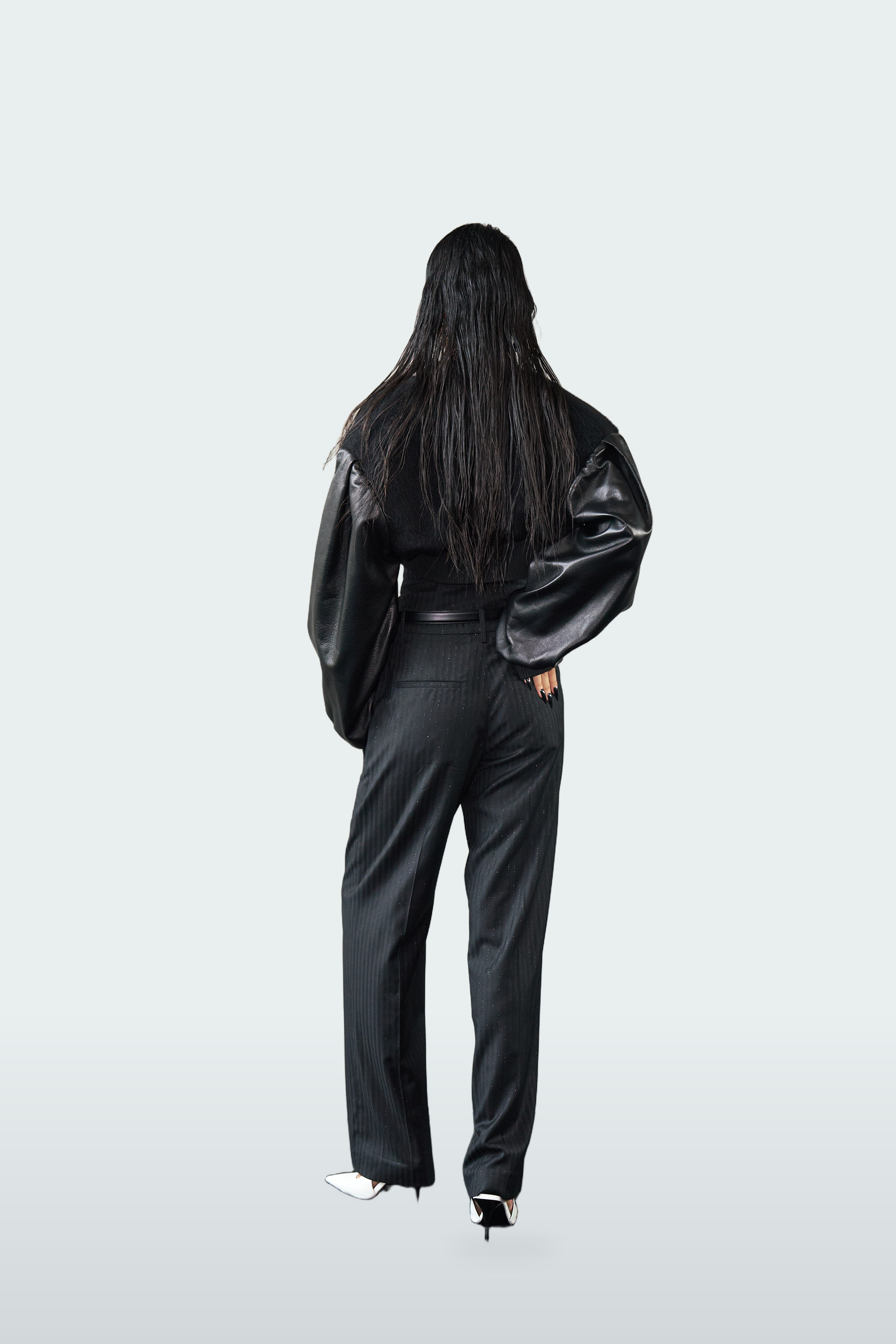 A model viewed from behind wearing a black outfit consisting of a shiny black jacket with voluminous sleeves and black wide-leg trousers, set against a light gray studio background.