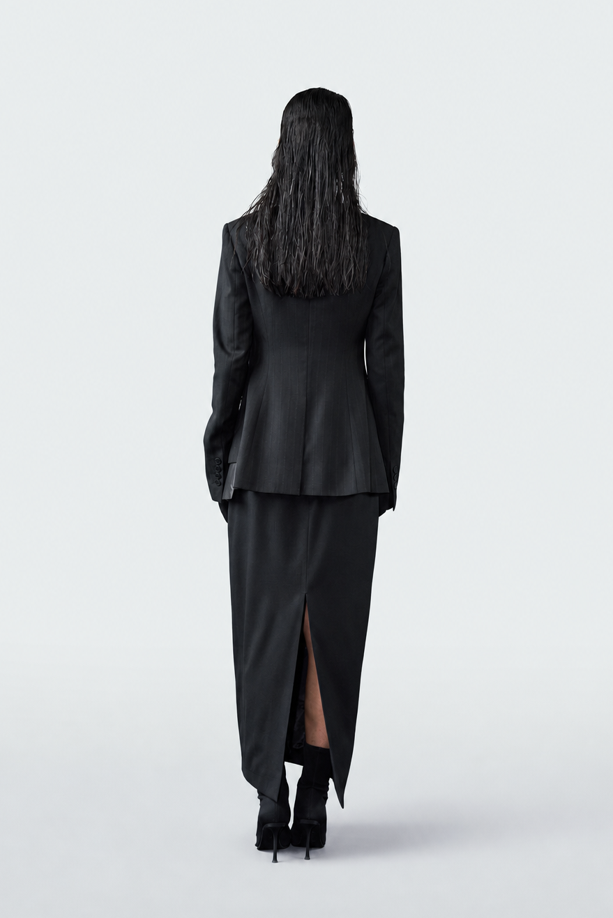 A model viewed from behind wearing a black long-sleeve blazer and a matching high-slit midi skirt, paired with black high heels, against a solid light gray background.