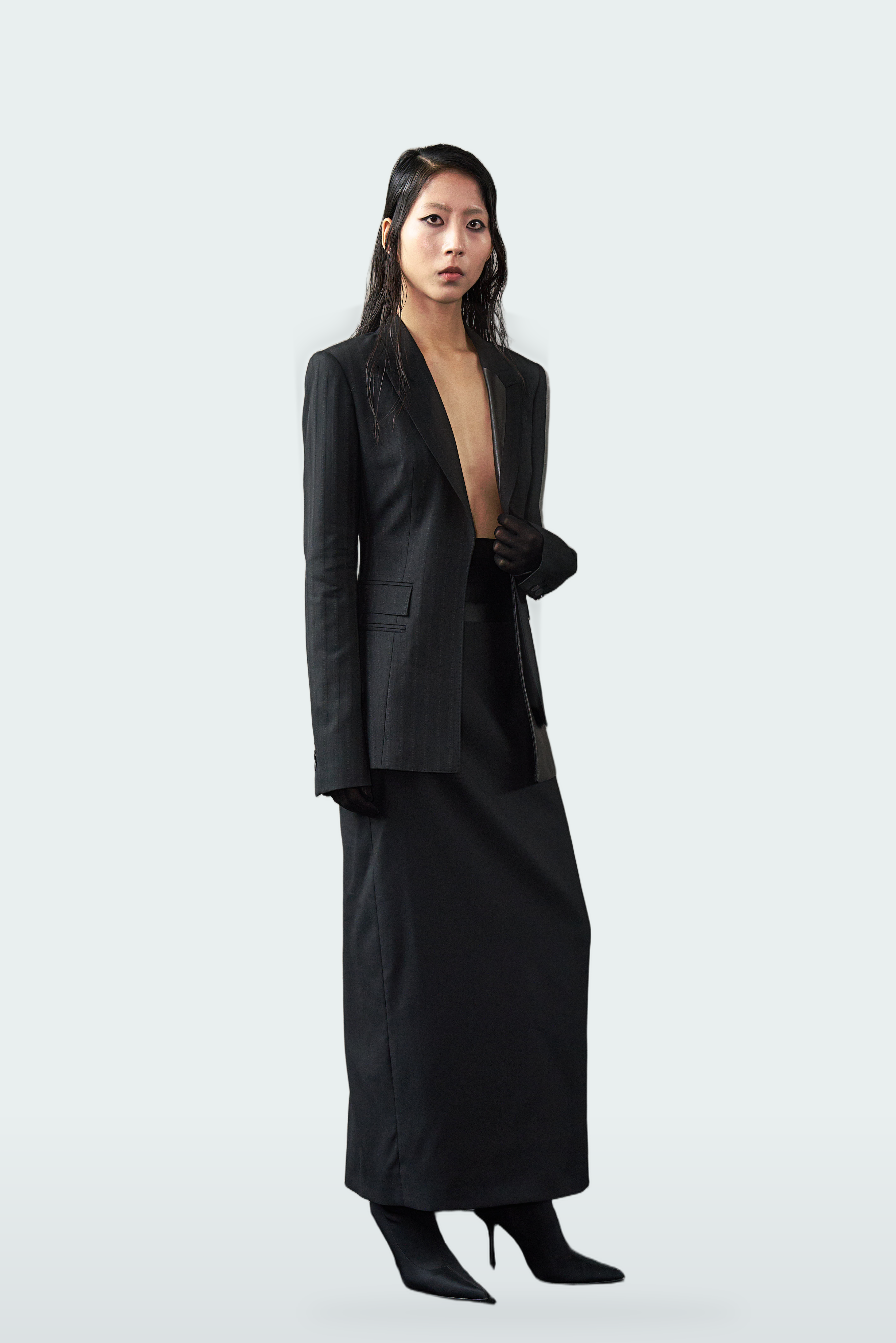 A model stands against a light gray studio background wearing a black tailored blazer with long sleeves, a matching long skirt, and black pointed heels.