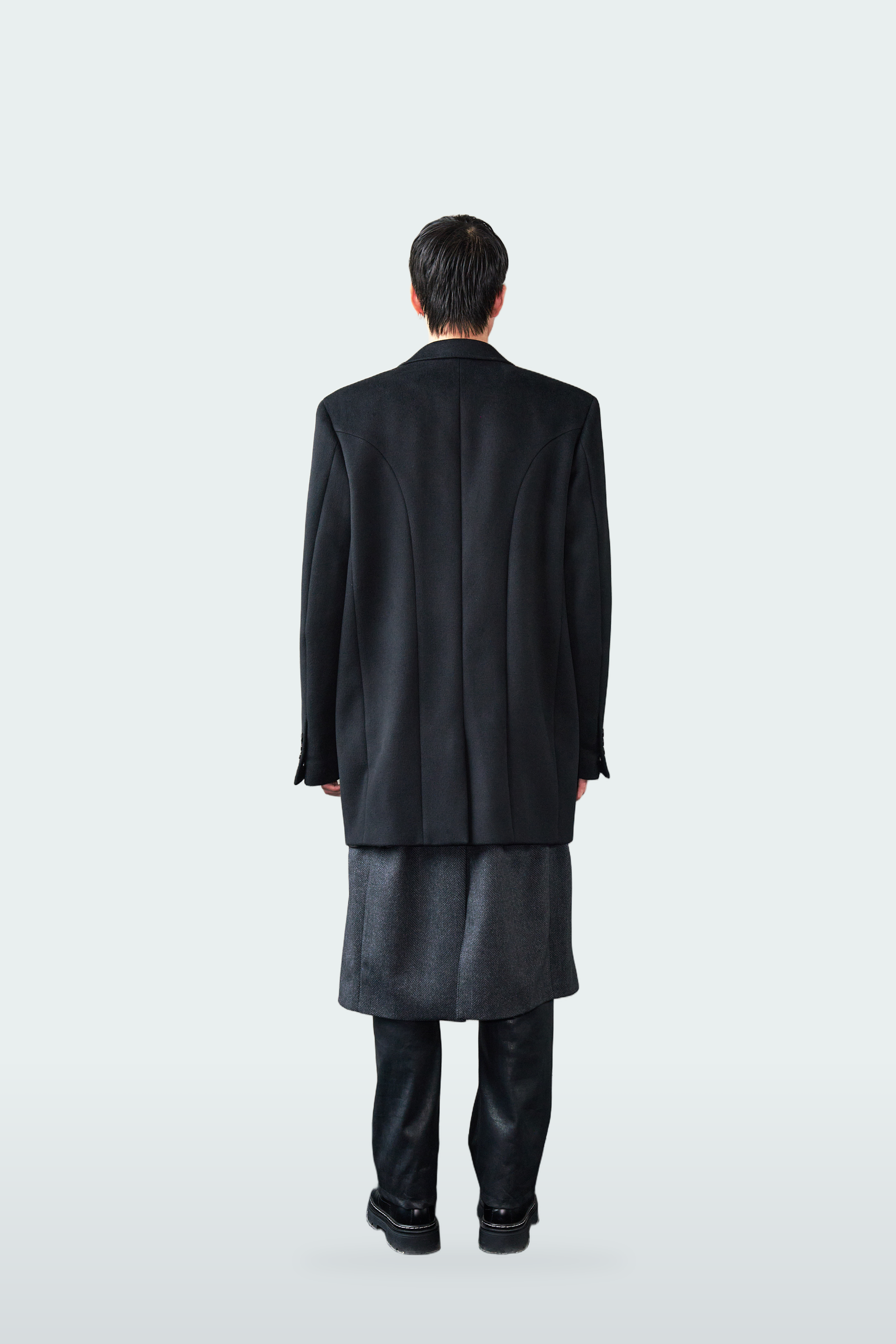 A model viewed from the back wearing a black longline overcoat, a dark gray textured skirt, black trousers, and black shoes against a light gray studio background.